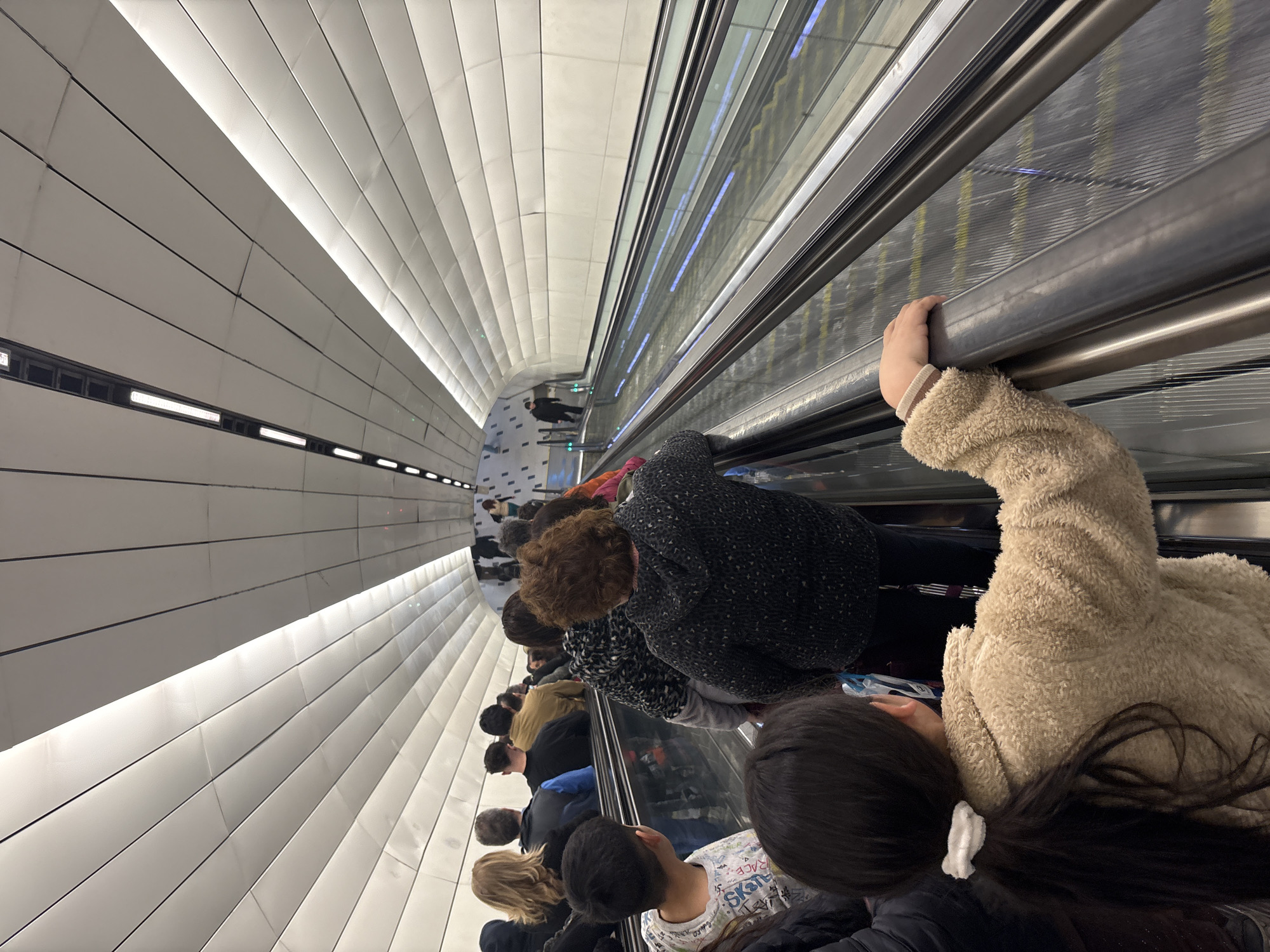 The image shows a group of people on an escalator in a subway station in Santiago, Chile. The escalator is moving upwards, and the people are facing forward, looking up the escalator. The station is modern and clean, with white tiled walls and ceiling. There are lights on the ceiling, and the escalator has a metal railing. The people are dressed in casual clothing, and most of them appear to be adults, though there are a few children visible. The image is taken from behind a young girl who is holding onto the escalator railing. The overall impression is one of a busy, functional public space.