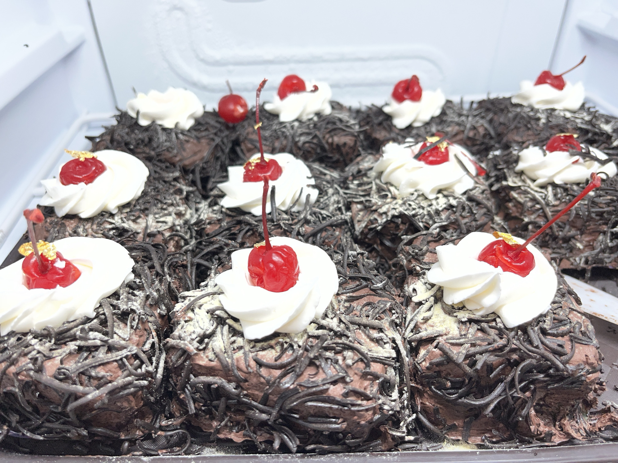 A close-up, eye-level, horizontal shot shows a tray of individually portioned chocolate cakes, possibly Black Forest cakes, arranged in a refrigerator. The cakes are covered in dark chocolate frosting and shredded chocolate, resembling dark strands. Each cake is topped with a dollop of white whipped cream, a bright red cherry, and a small piece of gold leaf. Several cherries are visible on stems, and some have fallen onto the cakes. The background is the white interior of a refrigerator. No people are present in the image, and no specific location in Lhokseumawe, Indonesia, can be identified.