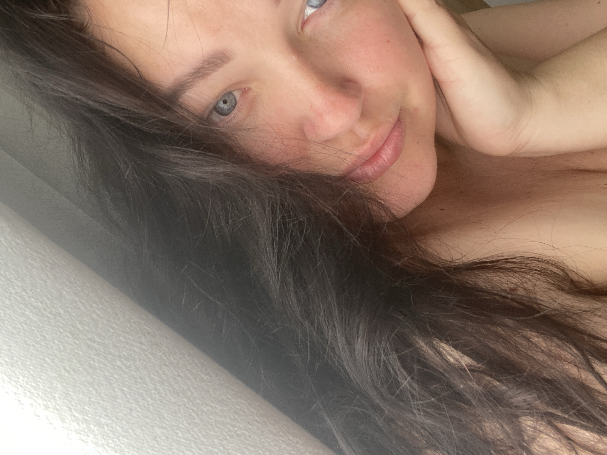 A close-up image captures a woman with fair skin and blue eyes. She rests her head against her hand, and her dark hair cascades across the image. A textured, light-colored surface is visible on the left edge, possibly a wall or bedding. The woman is positioned at an angle, with her face slightly tilted towards the camera. No other details or location information are discernible.