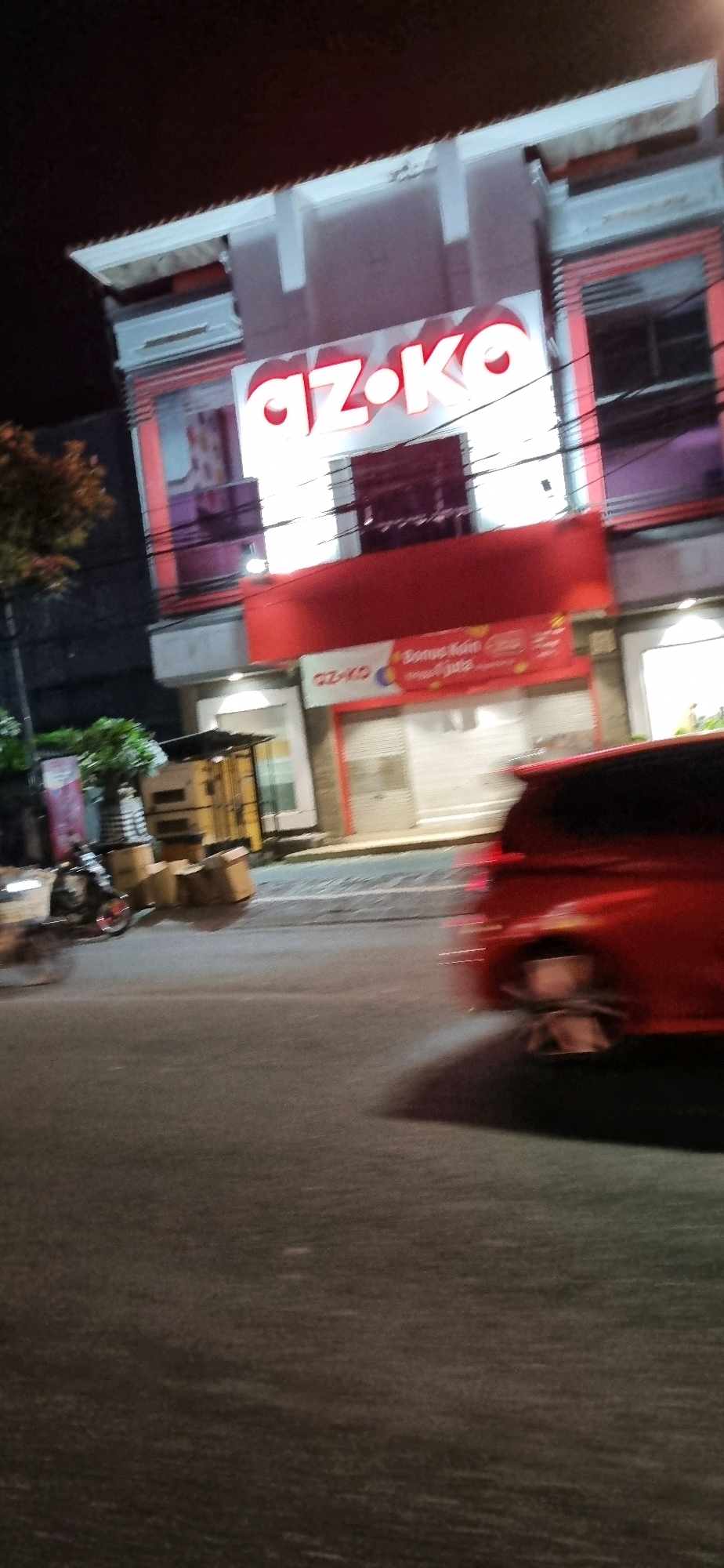 This is a low-light, motion-blurred, medium-shot photograph taken at night on a street in Denpasar, Indonesia. The primary subject is the facade of a two-story building with a prominent red and white sign displaying the illuminated text "AZ·KO" in large, white, sans-serif font with a red outline. The building appears to be a commercial establishment, possibly a retail store, as evidenced by large windows on the second floor and roll-down shutters on the ground floor. Below the "AZ·KO" sign, there is a red awning and a smaller banner that reads "AZ-KO" with an additional section that seems to advertise a promotion, with some text being illegible due to blur and low light.
To the left of the building, there is a motorcycle parked next to a small, open-air kiosk or stall with yellow metal framing and a corrugated metal roof. Several cardboard boxes are stacked near the motorcycle. A tree with dark foliage is visible behind the motorcycle and kiosk.
On the right side of the image, the rear end of a red car, likely a hatchback, is in motion, causing significant motion blur. The car's rear wheel and part of its bumper are visible. The street surface is dark asphalt, with some faint markings or reflections visible. The overall lighting is dim, typical of a street scene at night, with artificial illumination coming from the building's signage and possibly streetlights, which are not clearly visible. The scene suggests a commercial area that is either closing for the night or is in a period of low activity.