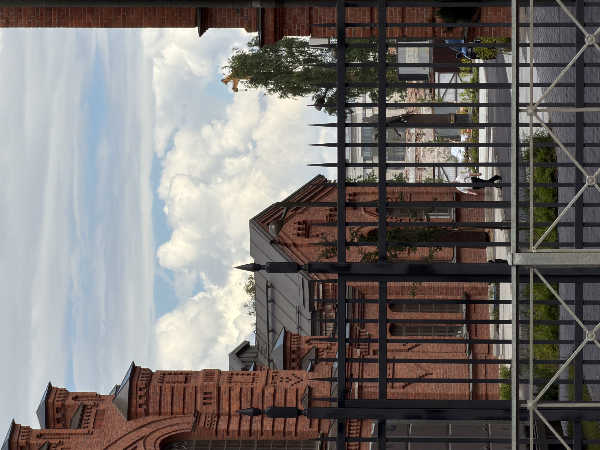 The image depicts an outdoor scene in Saint Petersburg, Russia. In the foreground, a black metal fence with pointed finials runs horizontally across the frame. Beyond the fence, a large brick building with a dark roof is visible. The building features arched windows and decorative brickwork. To the right of the main building, another brick structure with a dark roof and windows can be seen.
In the background, a tall green tree stands out against a cloudy sky. A yellow construction crane is visible above the tree. Further in the background, there are glimpses of other buildings and structures, including what appears to be a statue and a blue vehicle.
A single person, dressed in a white shirt and dark pants, is walking from left to right in the middle ground, behind the fence. The ground is paved with interlocking bricks. The overall impression is one of an urban environment, possibly a historical district or a construction site, with a mixture of old and new elements.