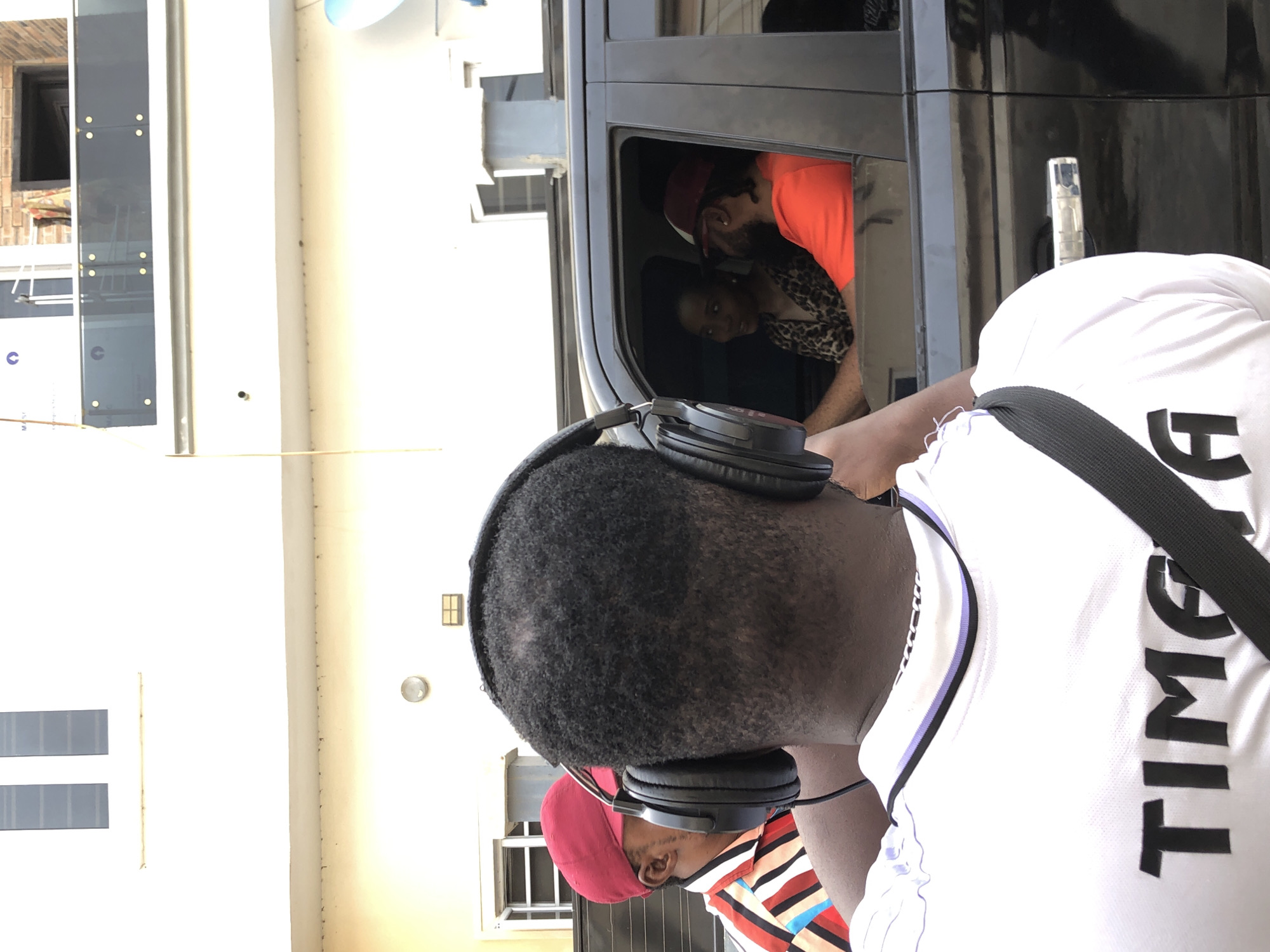 The image captures an outdoor scene in bright daylight, showing several individuals and a dark vehicle next to a building.
In the foreground, viewed from behind, is a person wearing black over-ear headphones. Their hair is short, dark, and curly. They are wearing a white short-sleeved shirt with bold black lettering on the back, which clearly reads "TIME". A black strap, likely from a bag, is visible over their right shoulder.
Through the open window of a dark-colored vehicle, positioned to the right of the foreground person, two individuals are visible. One is a man with a beard, wearing a red cap and an orange t-shirt, appearing to lean forward. Next to him, a woman with dark hair is visible, wearing a leopard print top. Both appear to be looking towards the foreground or slightly past them.
Partially visible in the lower-left, behind the main foreground subject, is another person wearing a red cap and a vertically striped multi-colored shirt.
The background consists of light-colored buildings with visible windows, suggesting an urban or suburban setting. The overall impression is a casual outdoor interaction or observation during the daytime.