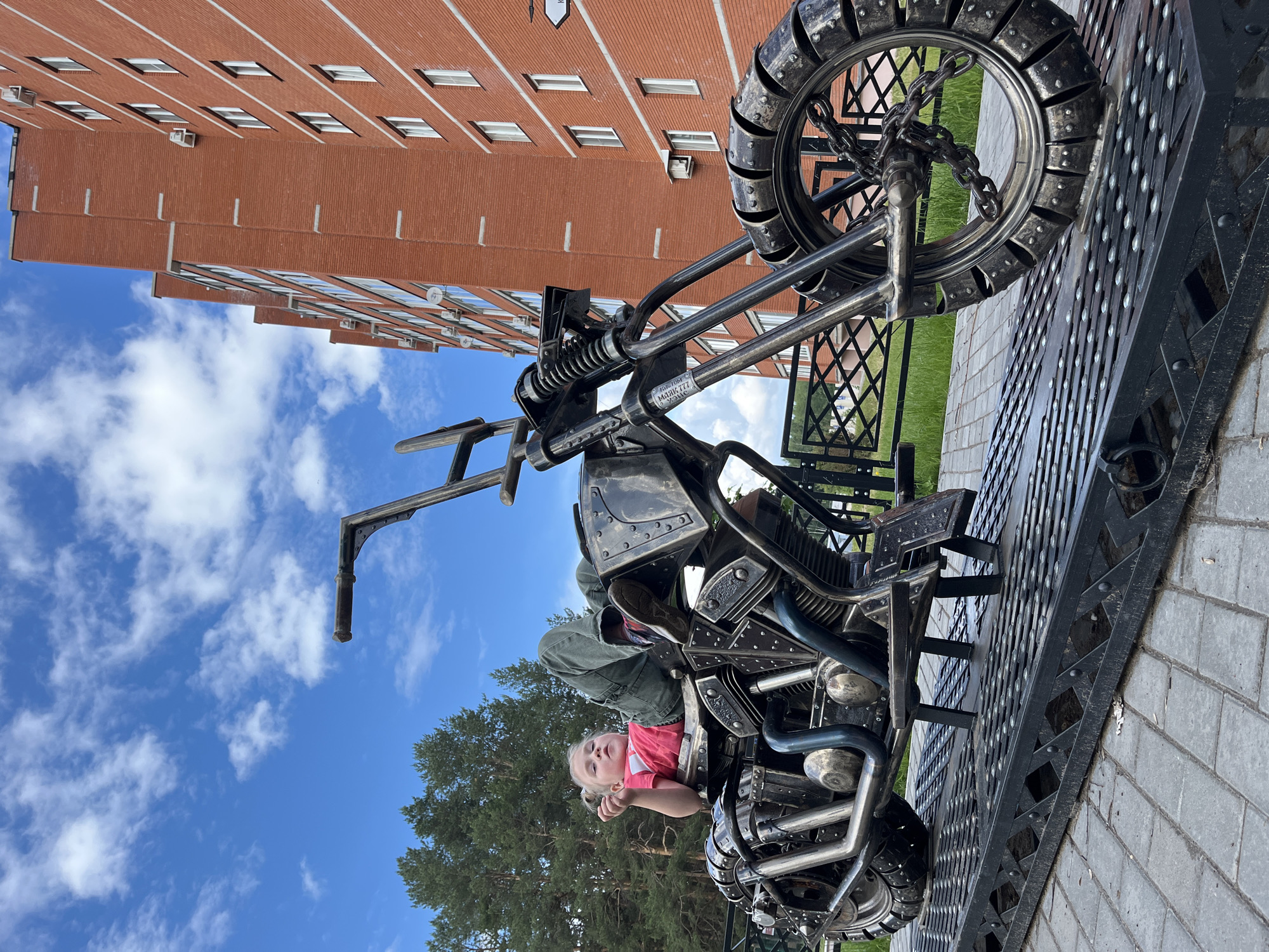 An outdoor scene features a large, intricately designed metal motorcycle sculpture, seemingly a public art piece or monument. The motorcycle is fabricated from dark metal with various metallic components, including visible springs, pipes, and a rugged, studded appearance on its body and wheels. The front wheel incorporates chains as part of its design.
A young girl with light hair, wearing a pink shirt and green pants, is seated on the motorcycle. Her left arm rests on the "fuel tank" area, and she looks slightly upwards with a calm expression.
In the background, a multi-story building constructed of red brick with numerous windows rises under a bright blue sky dotted with scattered white clouds. Green trees are visible between the building and the motorcycle. The motorcycle rests on a dark, textured metal platform, which itself is situated on a paved surface, likely a sidewalk or plaza. A metal fence is also partially visible on the right.
The scene depicts a sunny day. On a small silver plate affixed to the motorcycle's frame, the words "KUSTOM" and "МОЯК 777" are legible, along with "X-10" below them. The location suggested by the architecture and context is Chaikovsky, Russia.
