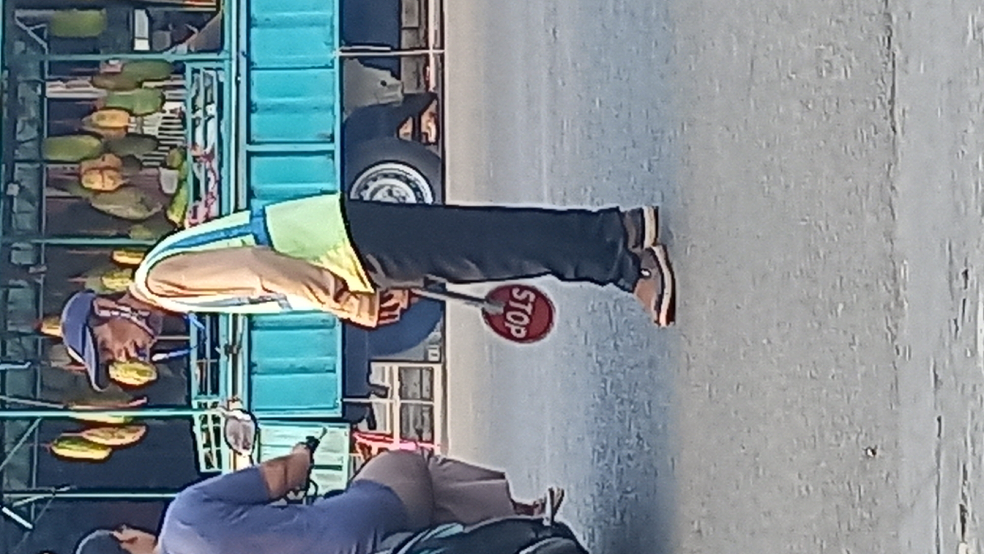 In Mangunsari, Indonesia, a man wearing a blue cap, a light brown long-sleeved shirt, and a yellow and blue reflective vest stands on a paved street, holding a red stop sign with white lettering. The sign is partially obscured and the lettering is reversed. He appears to be smoking a cigarette. In the background, a turquoise truck is parked, filled with durian fruits hanging from the top. To the left of the man, a person on a dark-colored motorcycle is partially visible, wearing a blue shirt. The ground is asphalt, and the lighting suggests it is daytime.