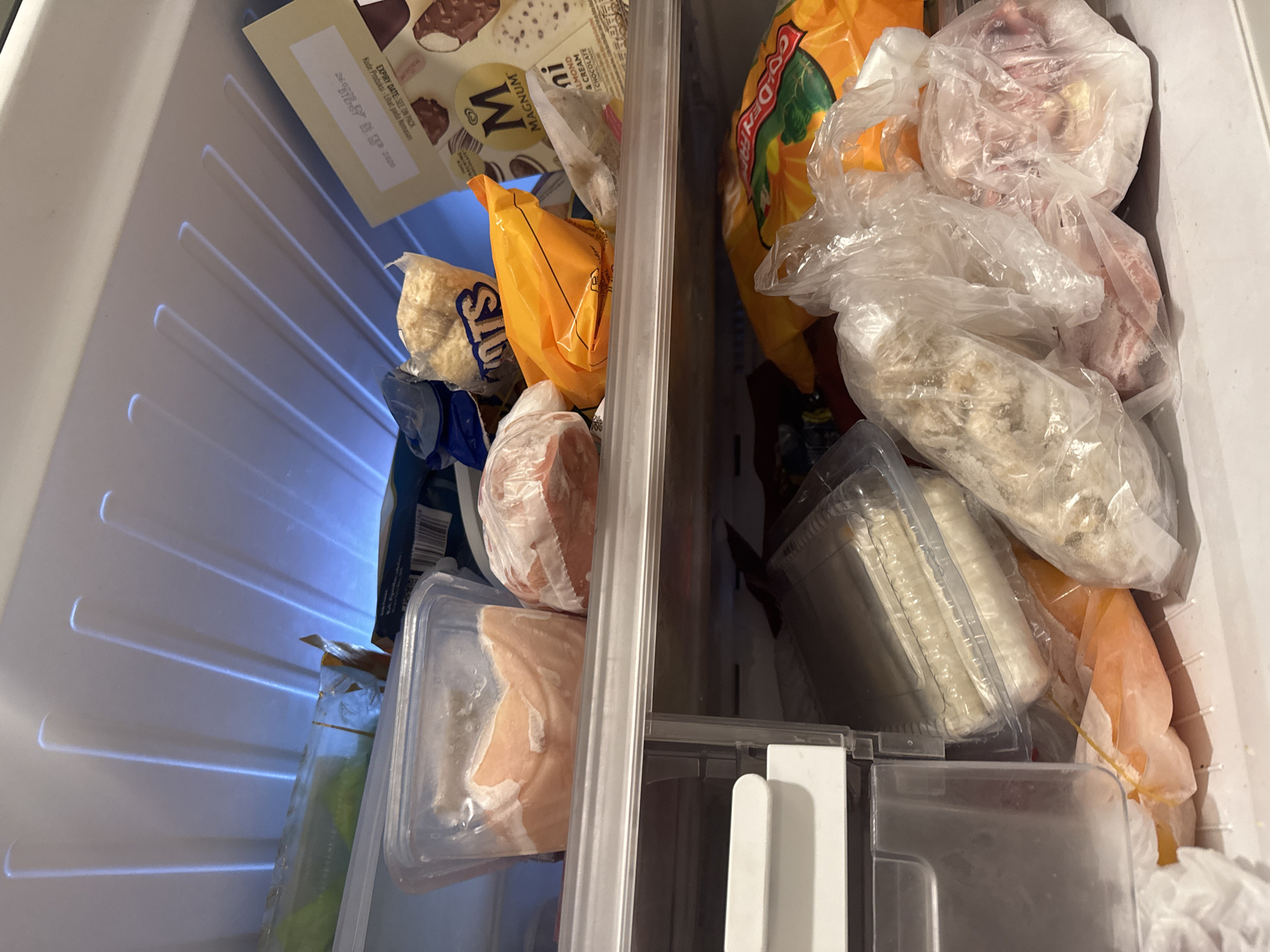 The image shows the interior of a freezer compartment, likely part of a refrigerator. The freezer is filled with various frozen food items. On the left side, there are frozen vegetables in a plastic container, a bag of frozen nuts, and what appears to be frozen meat. On the right side, there is a bag of Golden Raisins, and several plastic bags containing frozen items, possibly meat or fish. There is a divider running vertically down the center of the freezer compartment. In the top left is an open box of Magnum ice cream bars. Based on the packaging, the product originates in Bekasi, Indonesia.