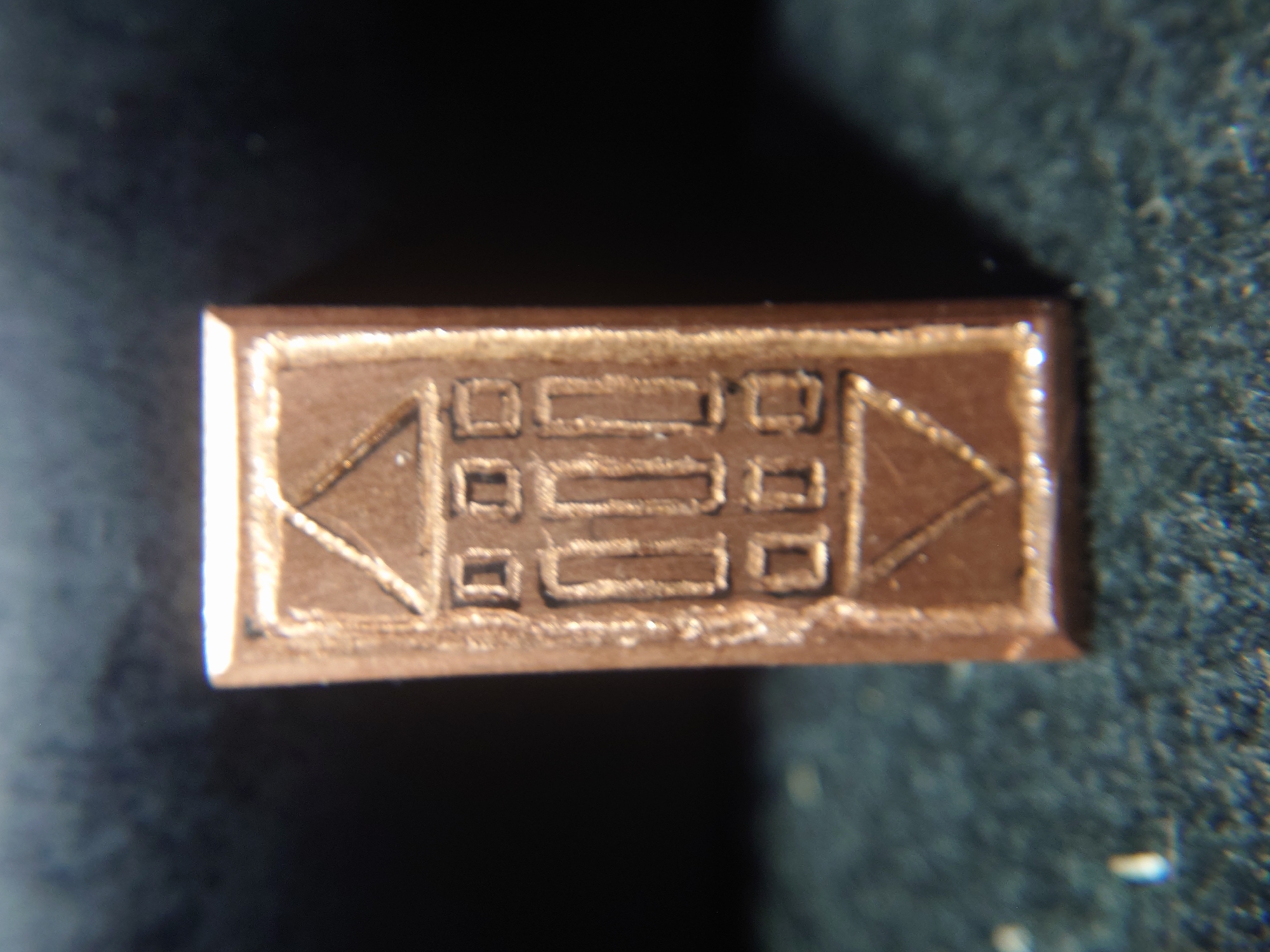 A rectangular copper object with a raised design sits on a dark, textured surface. The design on the object appears to be a stylized representation of a building or structure. The top of the object features a triangle within a square, with smaller squares and rectangles below it, arranged symmetrically. The bottom of the object has an inverted triangle. The lighting in the image highlights the metallic sheen of the object and casts a shadow to its right. The background is dark and out of focus. There is no discernible location information within the image that would suggest it is in Sokolohirsk, Ukraine.