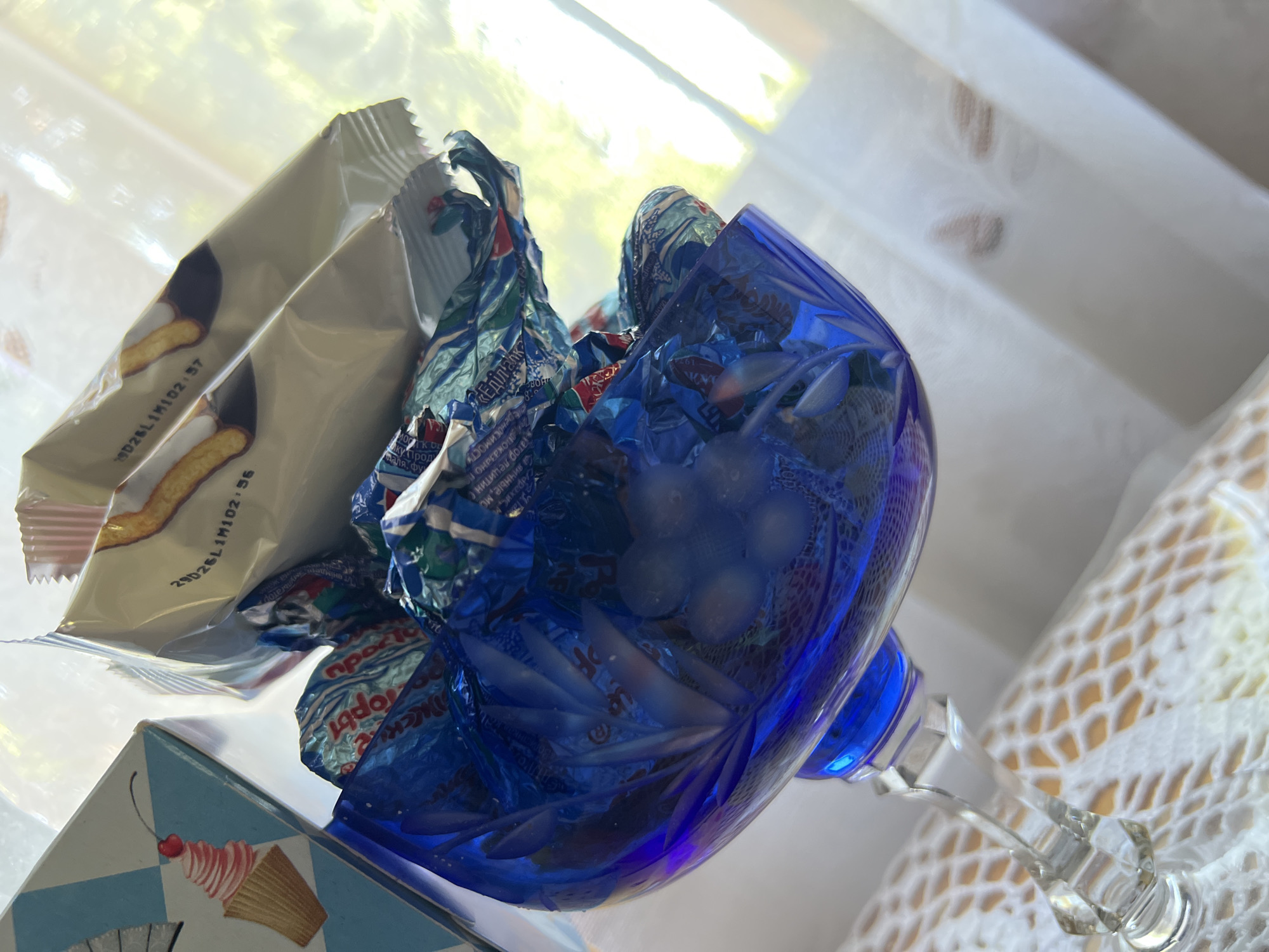 A close-up shot focuses on a blue crystal glass compote filled with individually wrapped candies. The candies are primarily blue, silver, and green, with some red accents. Above the compote, a partially open package of cookies is visible, with text that appears to be a date code "29026L1M102.56". To the left of the compote, a portion of a cardboard box with a graphic of an ice cream cone is visible. The background is out of focus, showing a bright, possibly windowed area with natural light and white curtains or fabric. The compote rests on a white crocheted doily. The image quality is slightly grainy. The scene appears to be indoors, possibly on a table, with soft lighting. No people are visible in the image. The location is identified as Khryashchevka, Russia.