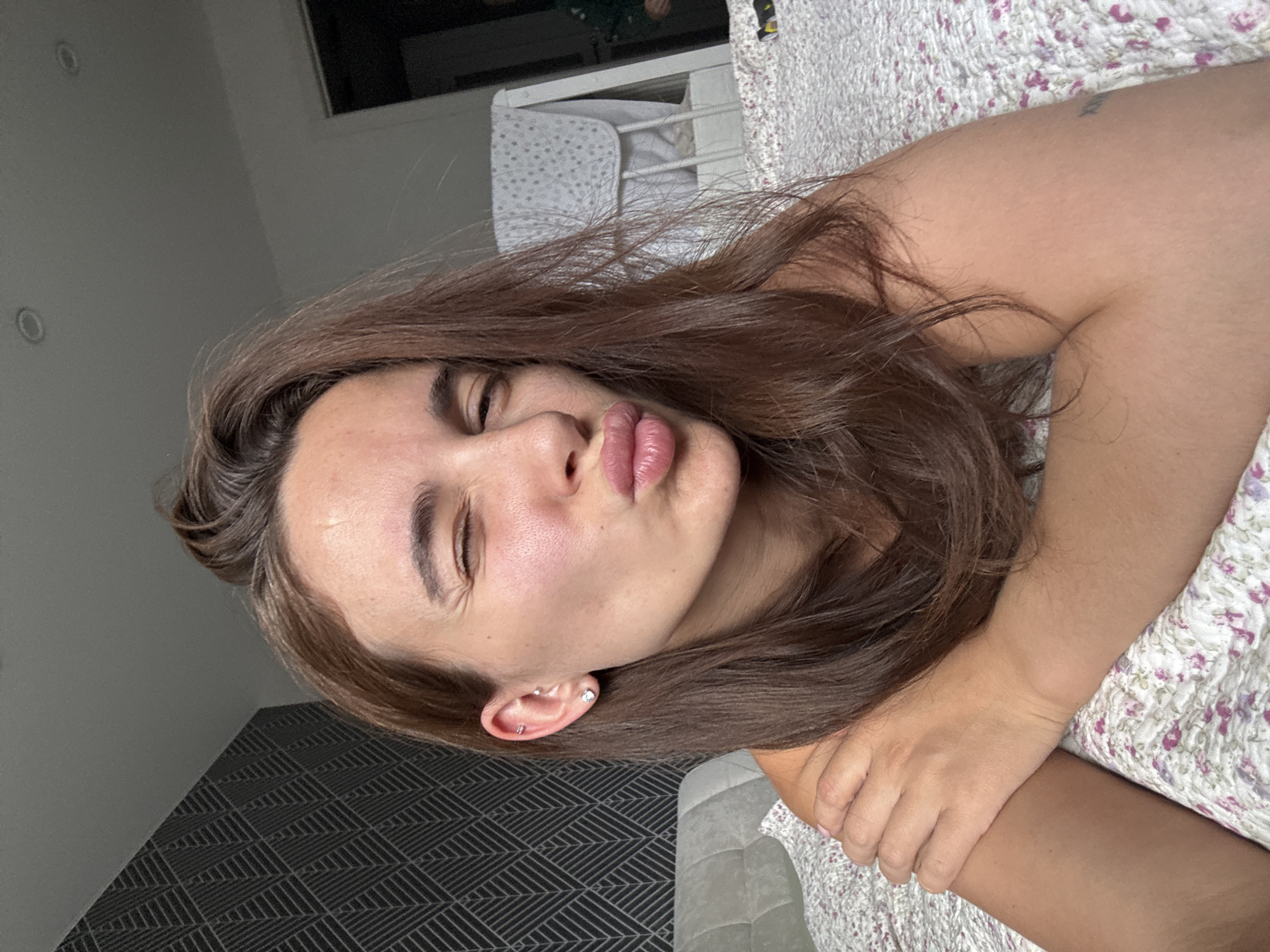 The image shows a close-up, self-taken photograph of a young woman lying on a bed, in what appears to be a bedroom. She has long, brown hair and is winking one eye while puckering her lips. She is wearing earrings and has a tattoo on her right shoulder. The bedspread has a floral pattern. In the background, there is a wall with a patterned wallpaper, a crib, and a window. The overall lighting suggests it is daytime. The location is identified as Chaikovsky, Russia.