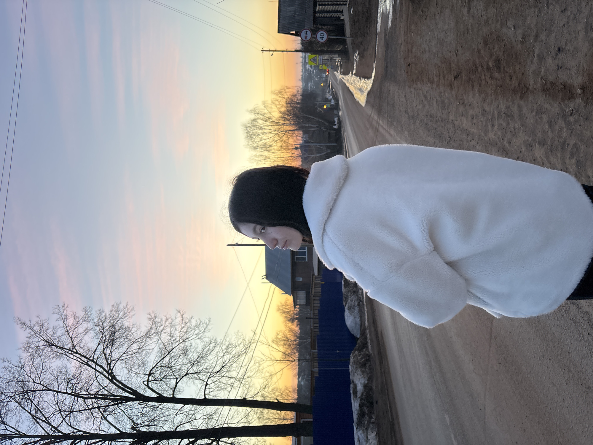 A person in a white jacket stands on a dirt road in Фоки, Russia, with a view of a colorful sunset. A house and a blue fence are visible in the background. A bare tree stands to the left, and overhead power lines are in view. There are road signs farther down the road. The sky is a mix of yellow, orange, pink, and blue.