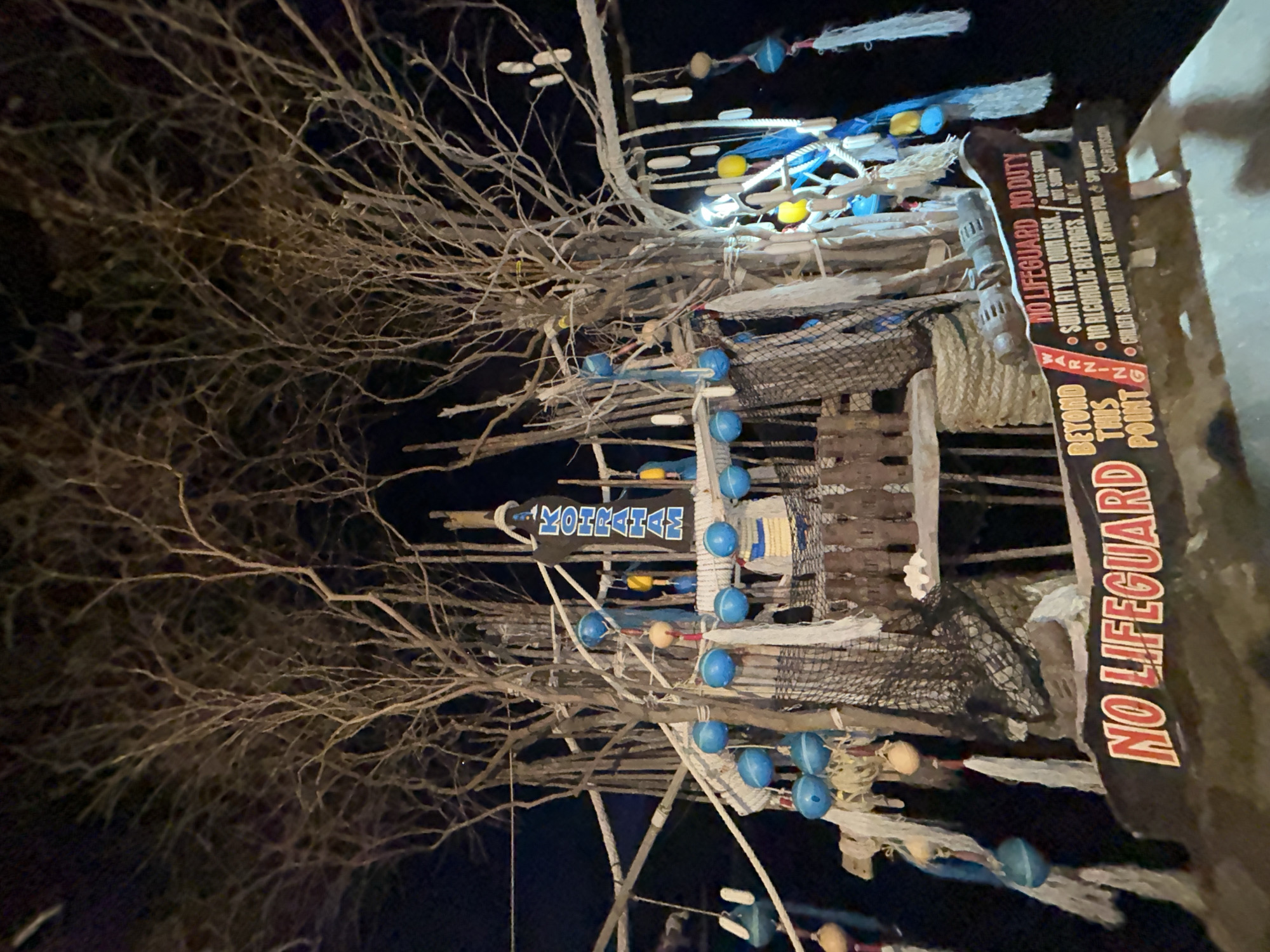 The image depicts an outdoor scene at night, possibly a bar or resort area, in Koh Phangan, Thailand. The central focus is a rustic, tree-like structure adorned with numerous blue and yellow buoys, fishing nets, and white tassels. At the top of the structure, a sign reads "KOHRRAHAA" in large blue letters. Below this, a wooden sign with a dark brown background and red and white lettering dominates the foreground. This sign features several warnings and disclaimers, including "NO LIFEGUARD," "SWIM AT YOUR OWN RISK," "NO ALCOHOLIC BEVERAGES," "CHILDREN SHOULD NOT USE THE SWIMMING POOL & SPA WITHOUT SUPERVISION," "ADULTS SHOULD NOT SWIM," and "BEYOND THIS POINT." The environment appears somewhat disheveled, with exposed tree branches forming the backdrop against the dark night sky. There are no people visible in the image.