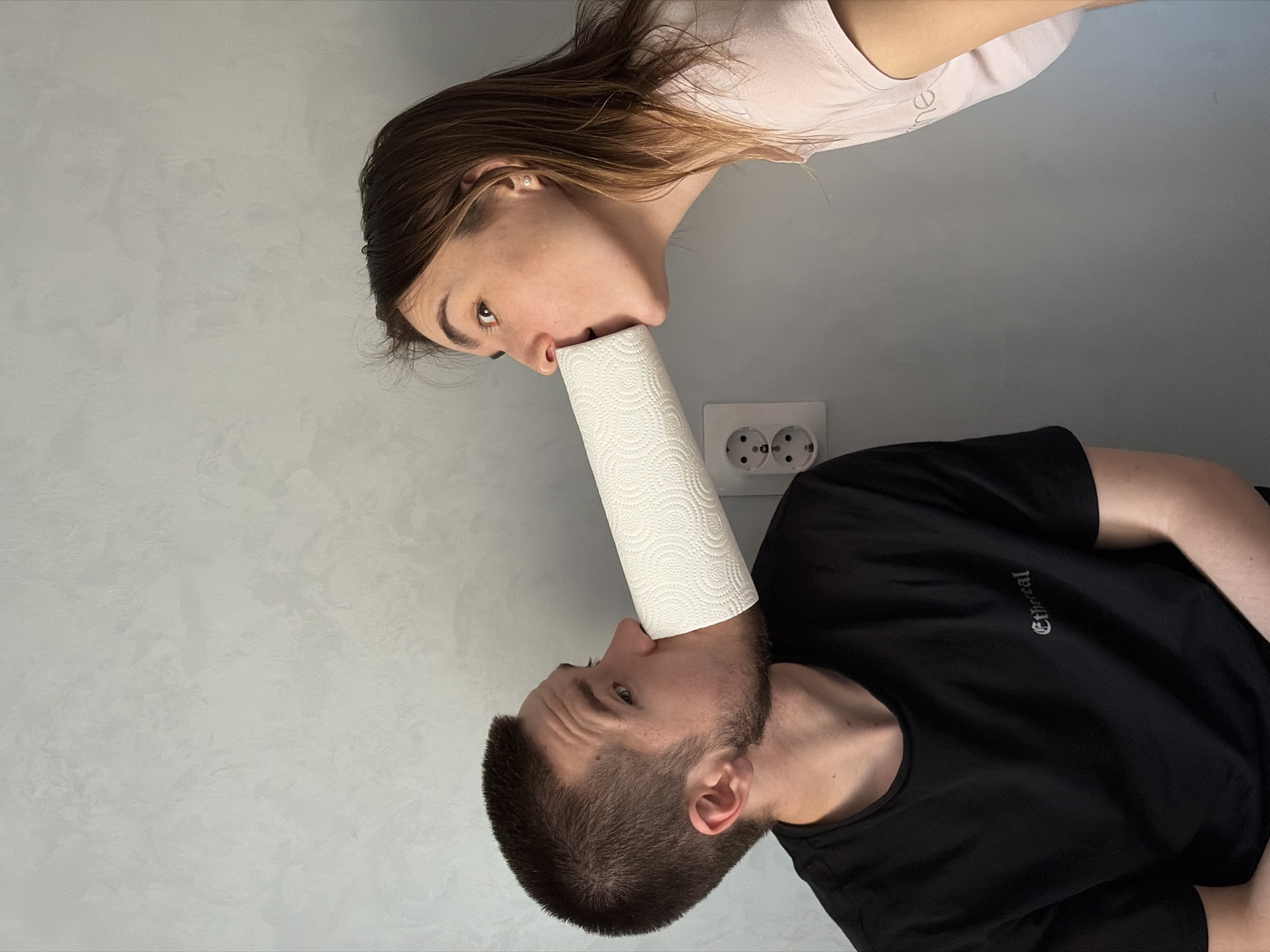 A light gray wall serves as the background for an image of two people oriented vertically, one above the other, appearing to connect by a roll of white paper towels. The person at the top, a fair-skinned female with long brown hair and wearing a light pink shirt, has the paper towel roll pressed against her nose. Beneath her, a fair-skinned male with short, dark hair and a beard is wearing a black t-shirt, and he also has the roll pressed against his mouth. A white electrical outlet with two circular openings is visible on the wall behind the roll of paper towels. The image orientation makes it appear as if the individuals are balancing the paper towel roll between them. The location is not evident in the image.