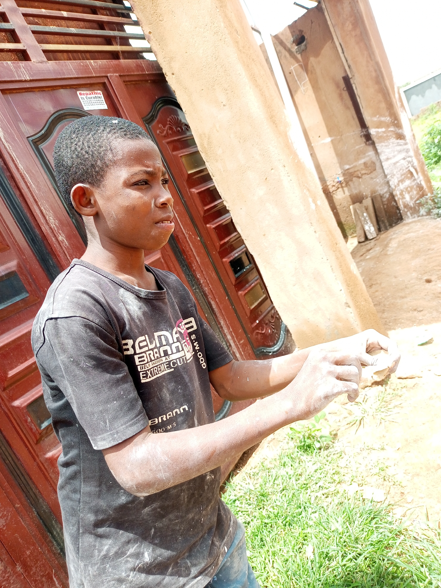 A young boy, appearing to be around 10 years old, stands in an outdoor setting with his upper body facing right. He is wearing a black t-shirt with some white dust or powder on it, and his arms are also dusted. The boy's hair is short and dark. He is looking to his right with a thoughtful expression, and his hands are positioned in front of him as if he is holding or manipulating something small.
The background includes a large, reddish-brown door or gate with horizontal slats and decorative elements. To the right of the boy, there is a light brown, roughly textured wall that appears to be part of a building. Further in the background, there are some loose stones or blocks, and beyond that, a glimpse of greenery and a lighter colored structure, possibly another building or fence. The ground appears to be a mixture of dirt and grass.
The scene suggests a casual, possibly domestic, environment. The dust on the boy and his t-shirt might indicate he has been involved in some sort of work or activity where dust is present, such as construction or crafting. The overall lighting indicates it is daytime.