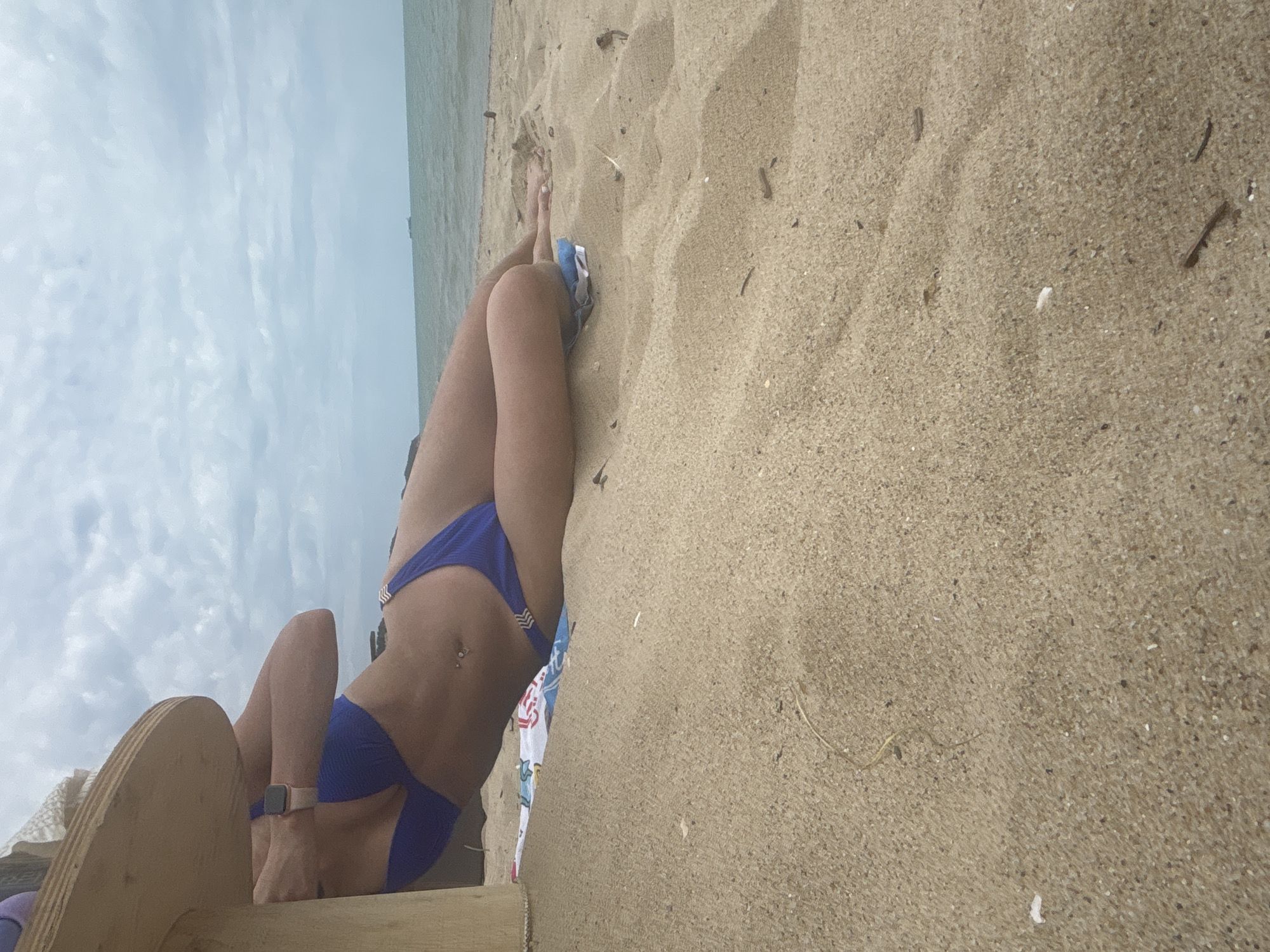 A person wearing a blue bikini is lying on the sand on a beach in Bliznatsi, Bulgaria. The person is partially leaning against a wooden structure. The ocean is visible in the background under a cloudy sky.