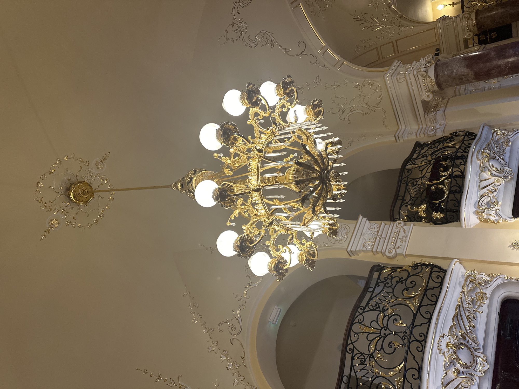 This media file captures a view looking upwards at an ornate, golden chandelier hanging from a cream-colored ceiling adorned with decorative plasterwork. The chandelier is the central object, featuring multiple light bulbs and intricate metalwork with crystal accents.
The ceiling is decorated with flowing, swirling patterns in a lighter shade and with gold accents, suggesting a grand interior space. To the right, a marble column with detailed white and gold decorative elements is visible, and part of a balcony railing with elaborate black and gold scrollwork is also present. The overall aesthetic points towards a luxurious and historical setting, possibly a theater, opera house, or grand hall.
The lighting suggests it is either daytime with the chandelier providing additional illumination, or evening. There are no people visible. The location cues, such as the architectural style, are consistent with the context of Odesa, Ukraine. The scene conveys a sense of opulence and elegance. No visible text is present in the image.