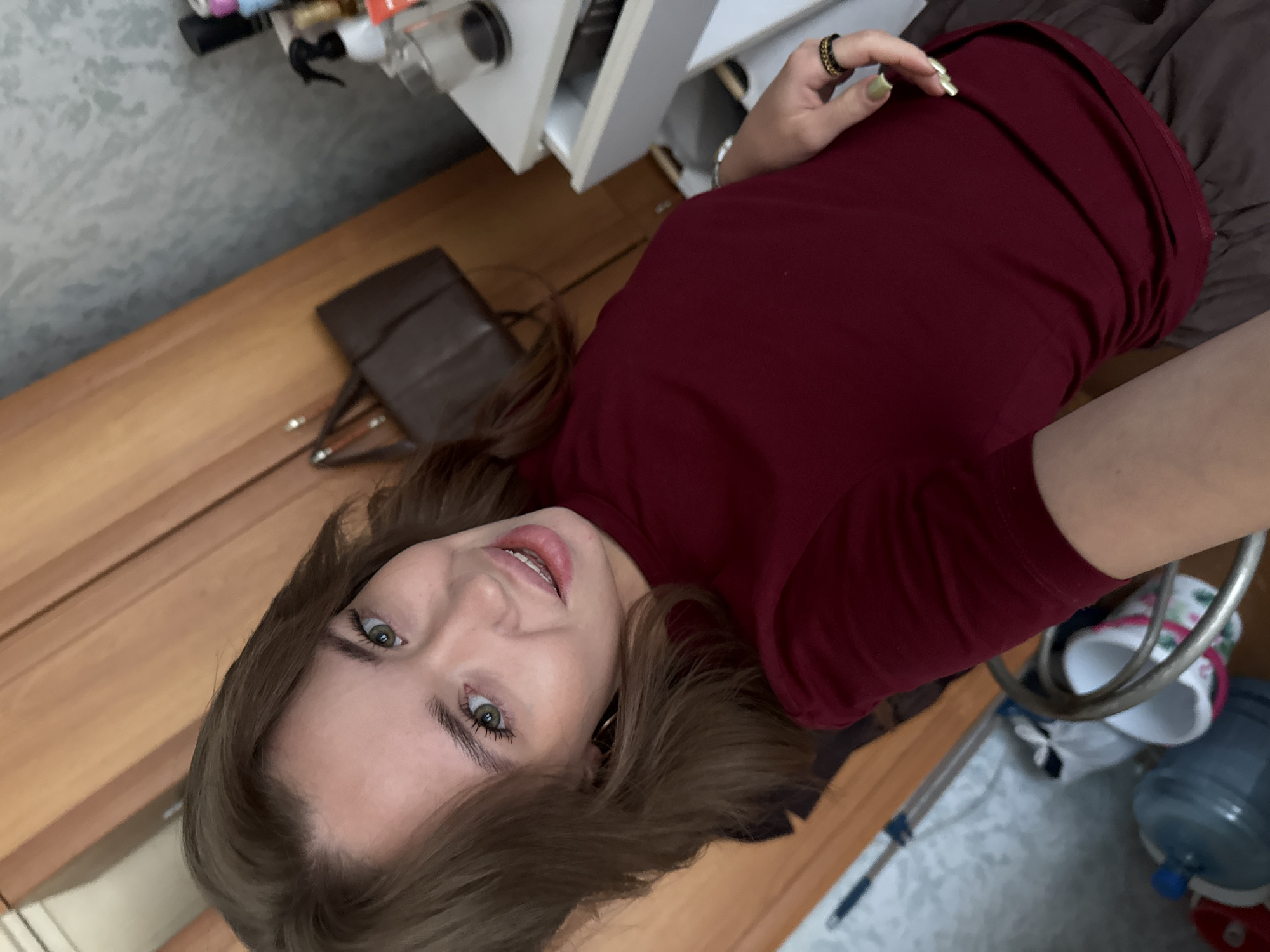 The image is a selfie taken indoors by a young woman with medium-length brown hair and green eyes. She is wearing a maroon t-shirt and dark grey skirt. The background appears to be a room, possibly a bedroom or living area, with a wooden wardrobe, a drawer unit with various items on top, and a floor cleaning tool in the lower left corner. The overall lighting suggests it is daytime, but the specific location within Chaikovsky, Russia, cannot be determined from the visual information provided.