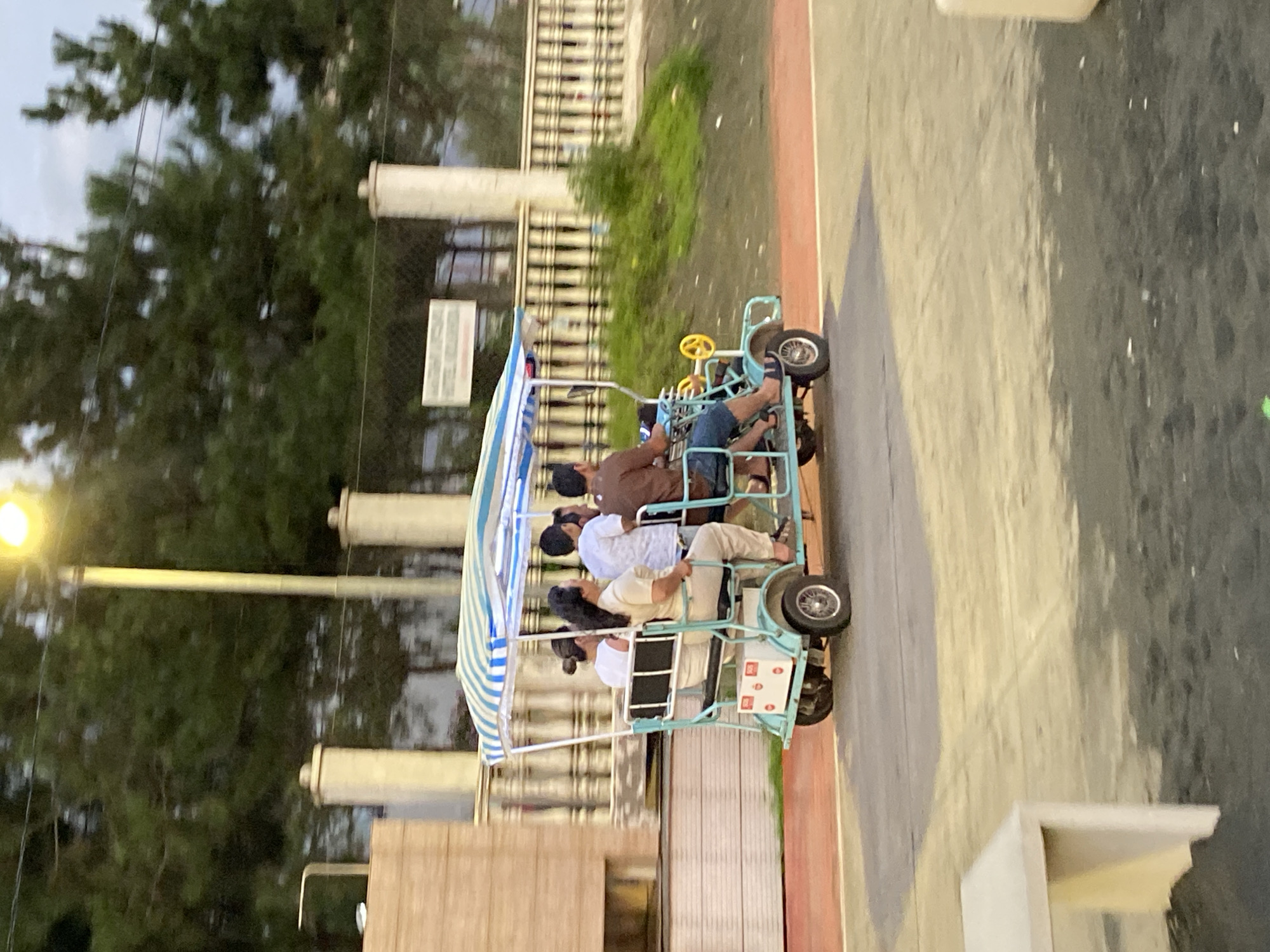 The image depicts a light blue, multi-person pedal cart with a striped canopy moving along a paved path. The cart is occupied by five individuals: a driver in the front operating the steering wheel and pedals, and four passengers seated behind. The passengers appear to be two women and two children. The setting is outdoors, with a line of coniferous trees visible in the background, a light pole with a yellow lamp, and a decorative fence. To the left, there is a light-colored building structure. The ground is a multi-colored paved surface, suggesting a park or recreational area. The overall scene suggests a casual outing in a public space, likely in Ureki, Georgia, a coastal resort town known for its magnetic black sand beaches and recreational facilities.
