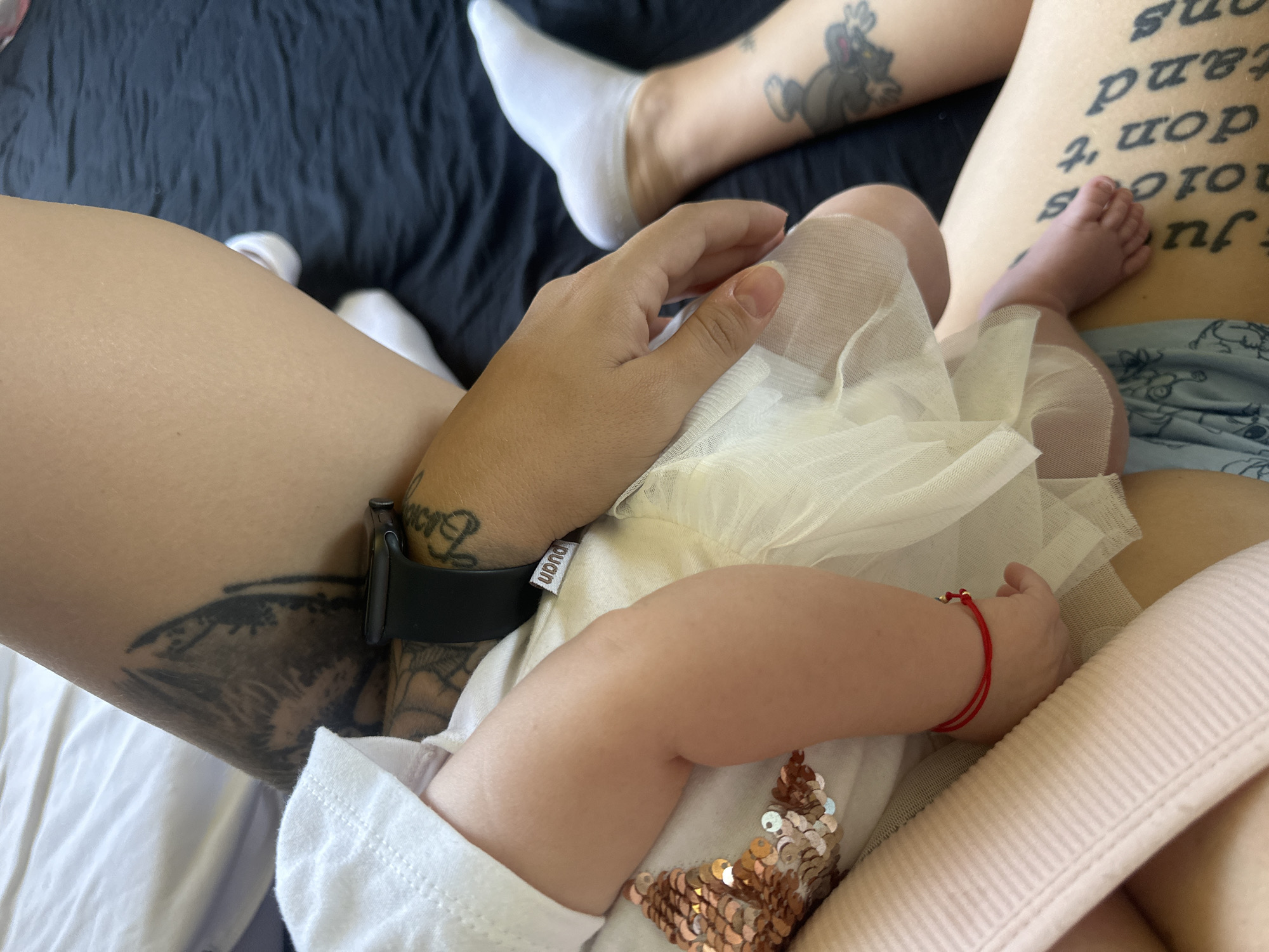 The image shows a close-up of a baby's arm and leg against the backdrop of a person lying on a bed with black bedding. The baby is wearing a white outfit adorned with sequined starfish and a sheer white tutu. A thin red bracelet with a gold clasp is visible on the baby's wrist. The person on the bed has multiple tattoos, including a large one on their thigh and ankle, and is wearing white socks. An Apple Watch is visible on the person's wrist. The lighting suggests an indoor environment, possibly a bedroom. The image does not provide any specific information or visual cues that would allow for the determination of its location in Plovdiv, Bulgaria.