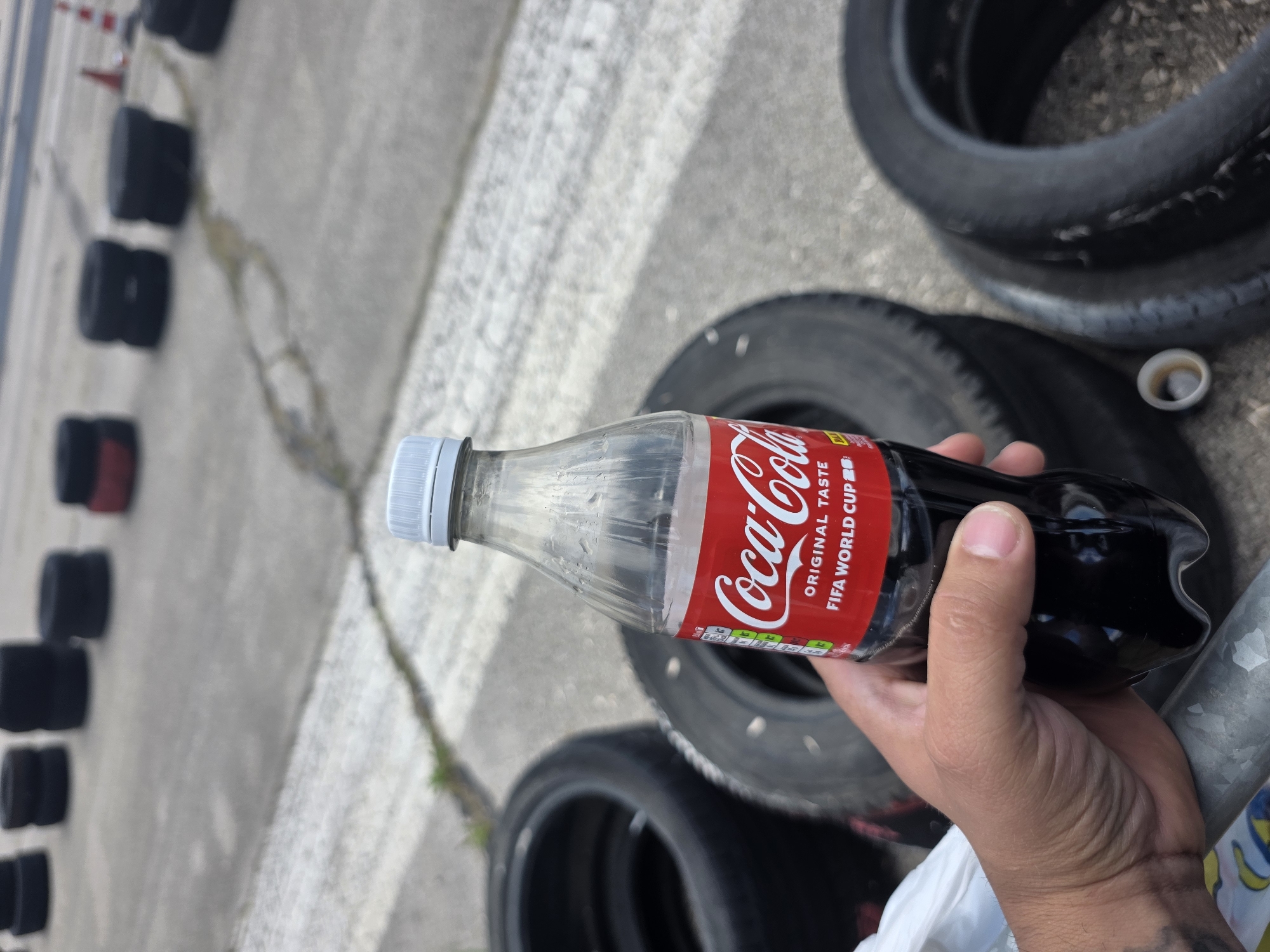 A close-up shot shows a hand holding a plastic bottle of Coca-Cola with a red label. The bottle is partially filled with a dark liquid. In the background, stacks of black tires are arranged on a gray paved surface, possibly a race track or a go-kart track. The scene is outdoors, with a slightly overcast sky. The background shows further stacks of tires and a few orange traffic cones. The location is identified as Akatsievo, Bulgaria.