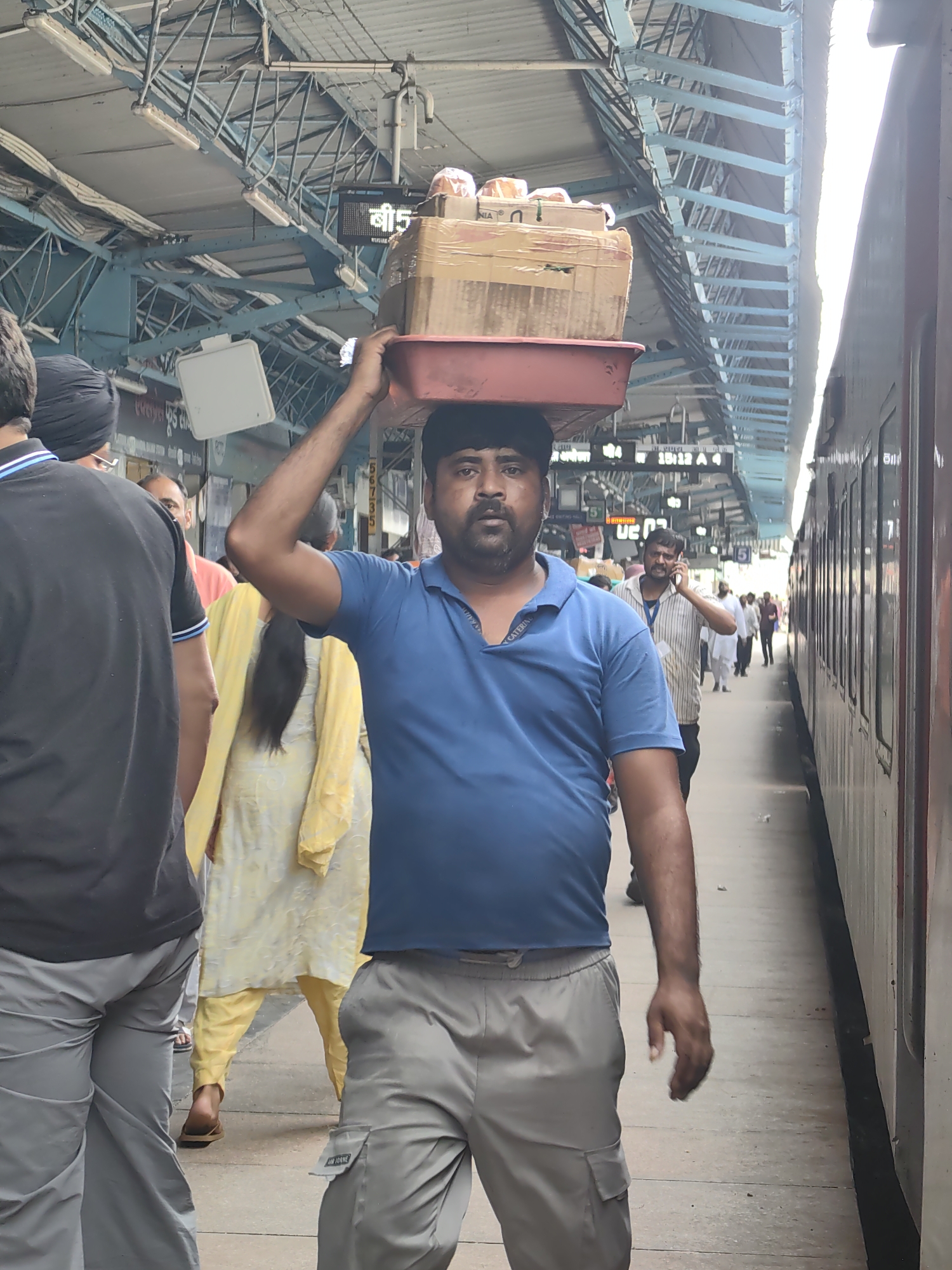 A man, likely a porter, walks forward carrying a stack of boxes on his head at the railway station in Khandwa, India. He is wearing a blue t-shirt and grey cargo pants. The boxes appear to be cardboard and are stacked on a red tray. In the background, other people can be seen on the platform, and a train is parked next to the platform. The scene takes place under the covered roof of the railway station, with metal support structures visible.