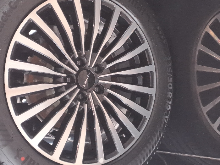 A close-up shot showcases the intricate design of a car wheel. The wheel features numerous spokes, alternately shaded in dark gray and silver. A black hub displays the inscription "TENST". The tire, marked with "235/50 R19 V", is partially visible. Another similar wheel appears blurred in the right edge of the image. The focus is sharp on the primary wheel, highlighting its details and materials.