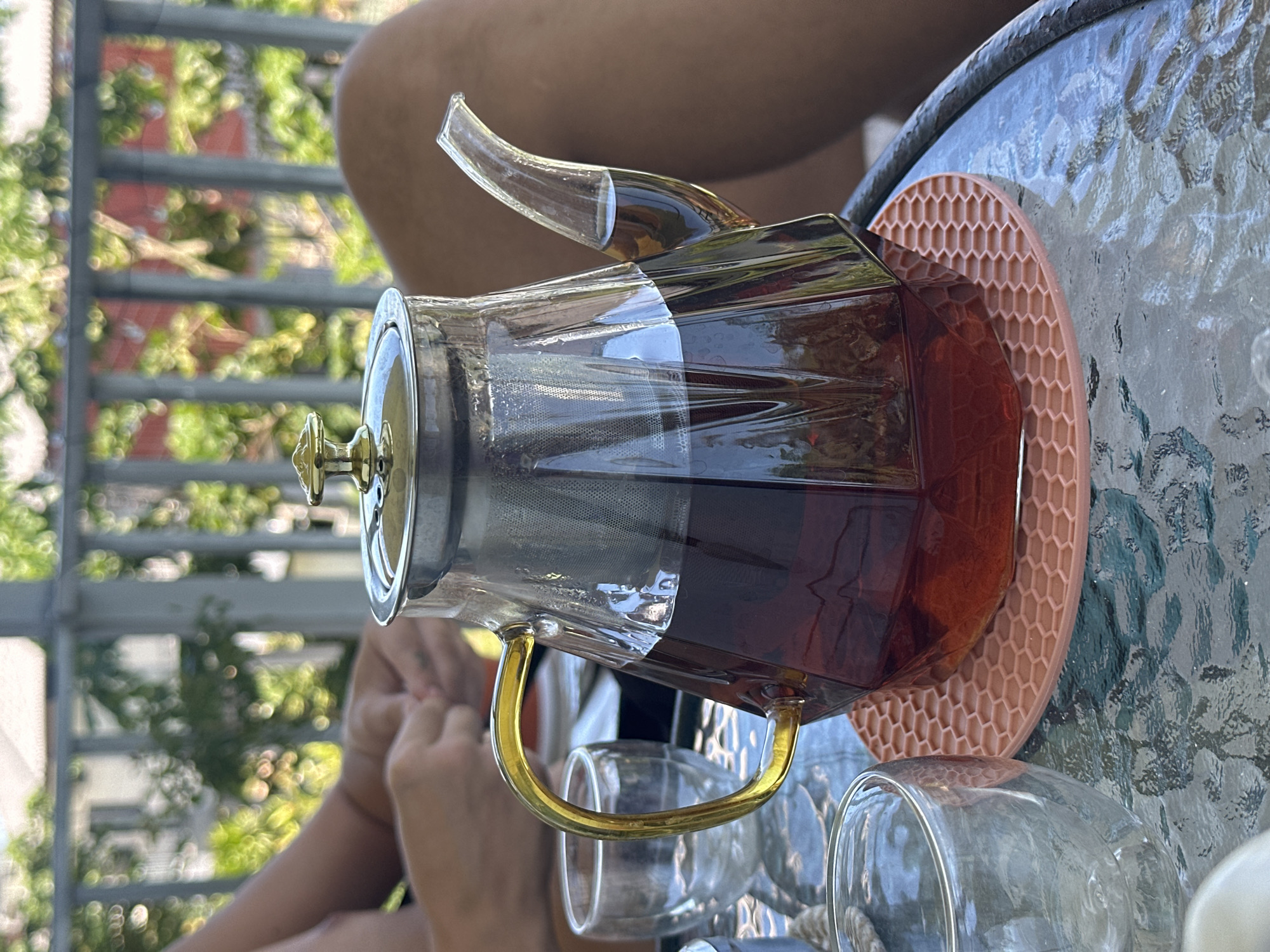 In Ko Samui District, Thailand, a clear glass teapot filled with dark amber liquid sits on a patterned pink coaster on a glass-topped table. The teapot has a metallic lid with a gold handle and a gold handle on the side. A metal infuser is visible inside the teapot. Two clear glasses, one partially filled with ice, are placed next to the teapot. In the background, a person's arms and legs are visible, suggesting they are sitting at the table. The scene appears to be on an outdoor porch or balcony, with a railing and lush green foliage visible beyond. The lighting suggests it is daytime, possibly late morning or early afternoon.