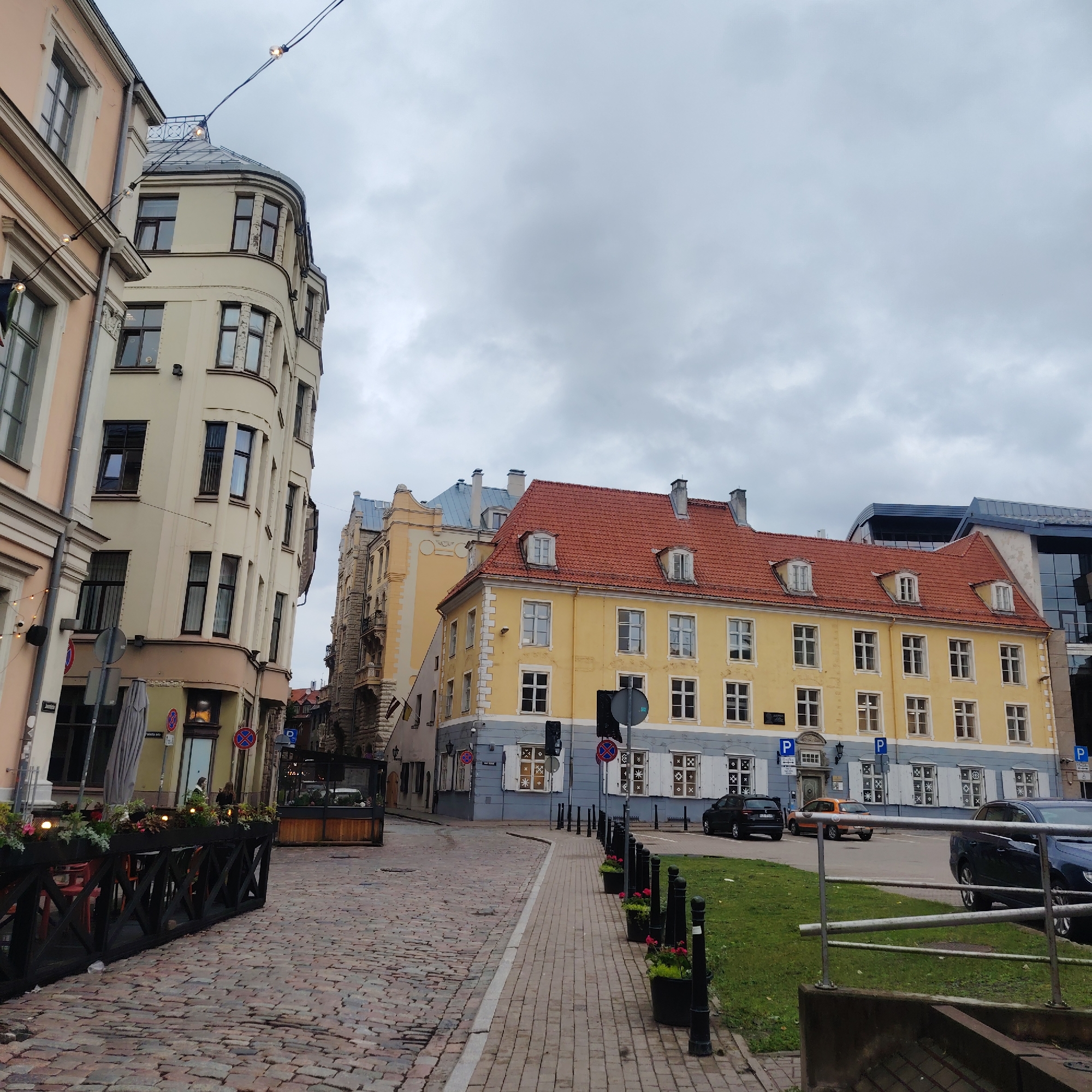 The image depicts a street scene in Riga, Latvia, on an overcast day. In the foreground, a cobblestone street curves to the right, bordered by a paved sidewalk. Along the sidewalk, a series of black bollards are interspersed with planters containing red flowers and greenery. Black wooden fencing with a crisscross pattern encloses an outdoor seating area with tables and chairs, likely for a cafe or restaurant.
Several buildings line the street. On the left, a pale pink building with multi-paned windows is partially visible. Adjacent to it is a cream-colored building with a distinctive curved facade and numerous windows with dark frames. Further down the street, on the right side, is a bright yellow building with a red tiled roof. This building features dormer windows and a row of white-shuttered windows on its lower level. Behind the yellow building, a more modern structure with dark glass and a grey roof is visible.
Traffic signs are present, including blue parking signs with a white 'P'. Several cars are parked or driving on the street. A dark-colored car is parked near the yellow building, and an orange car is further down. A dark blue car is parked in the right foreground. String lights with exposed bulbs are strung across the upper portion of the image, indicating a festive or decorative element. The sky is predominantly grey and cloudy. The scene appears to be a commercial or residential area in an older part of the city.