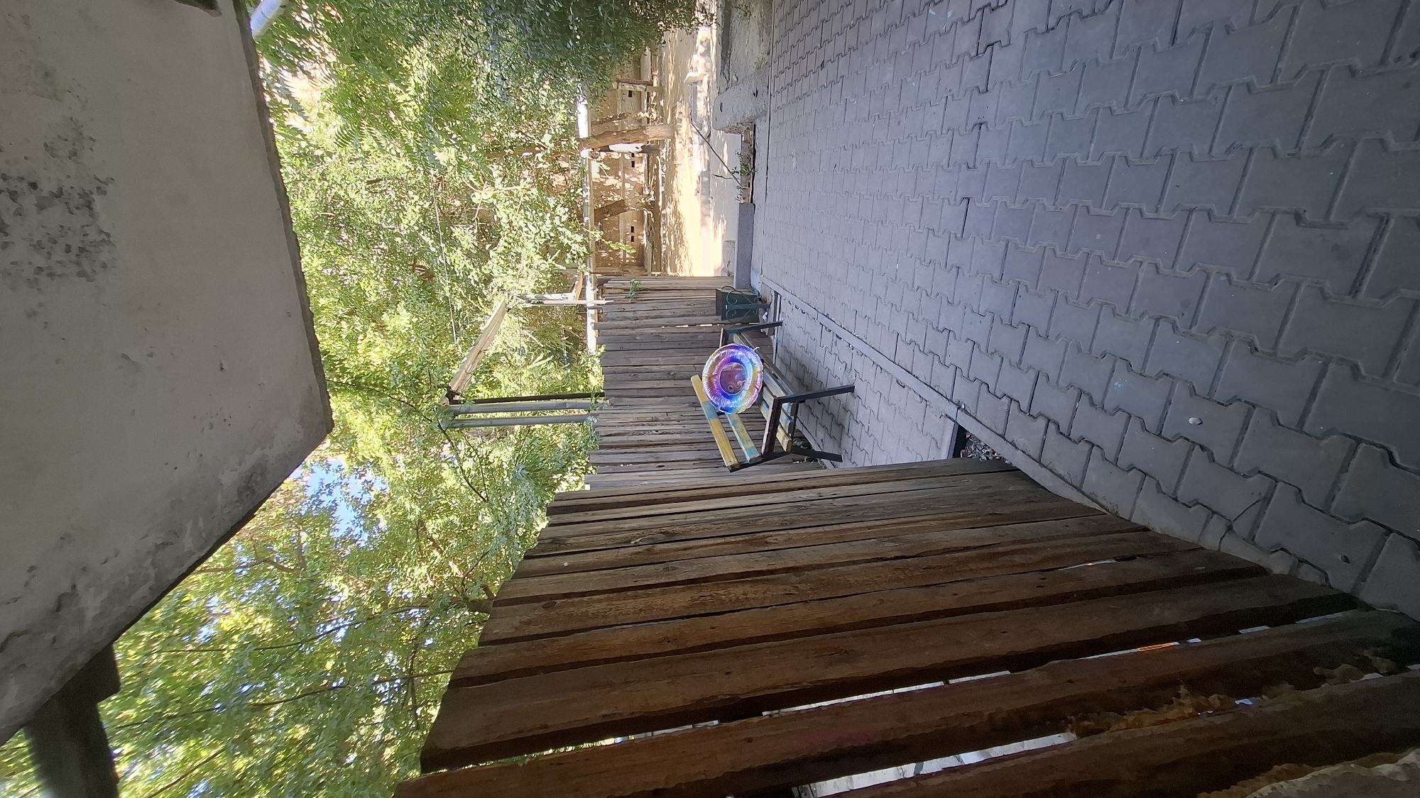 The image captures an outdoor scene in Shymkent, Kazakhstan. A wooden fence dominates the left foreground, its weathered planks running vertically. To the right of the fence, a paved walkway made of interlocking gray pavers extends into the background. A bench with a colorful, circular object resembling a balloon or a bubble rests on it. The bench appears to be made of wood with metal legs. Behind the bench, a green trash can is visible. Trees with lush green foliage form a backdrop, suggesting a park-like or courtyard setting. The scene is illuminated by natural daylight. In the far background, there are hints of structures and more trees, suggesting this is an outdoor public space.