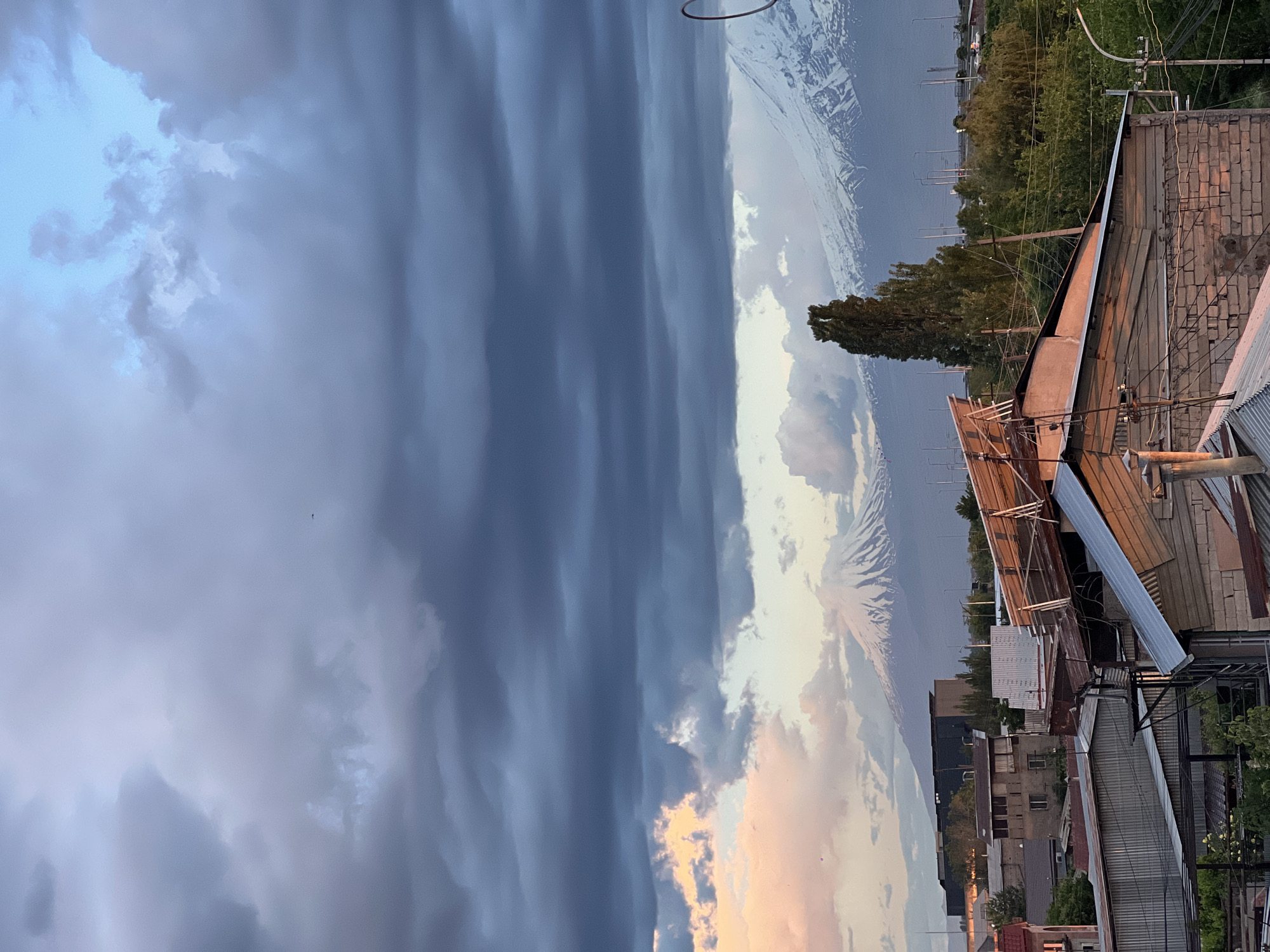 The image depicts a cityscape in Yerevan, Armenia under a dramatic sky. Dark, striated clouds dominate the left side of the frame, contrasting with brighter, sunset-tinged clouds near the center. In the background, the snow-capped peak of Mount Ararat is visible. In the foreground, a cluster of rooftops, utility poles, and trees fill the right side. The scene is shot from an elevated position, overlooking the city. Buildings of various heights and architectural styles are visible. No people are visible.