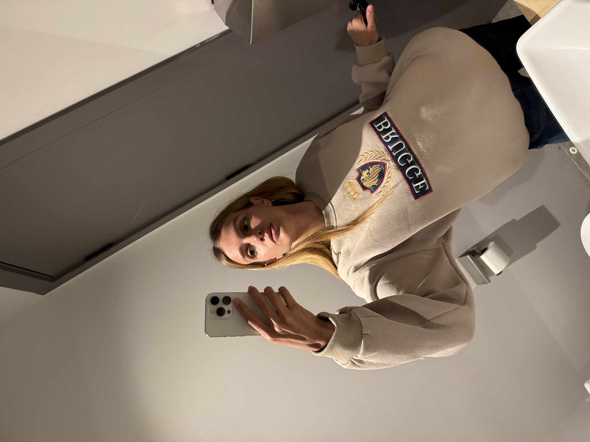 A blonde woman is taking a selfie in a bathroom. She is wearing a beige sweatshirt with the word "BKNCCE Belgium" embroidered on it. She holds a white iPhone in her left hand. A toilet paper holder is on the wall. The bathroom is likely located in Tbilisi, Georgia, based on the embroidered sweatshirt.