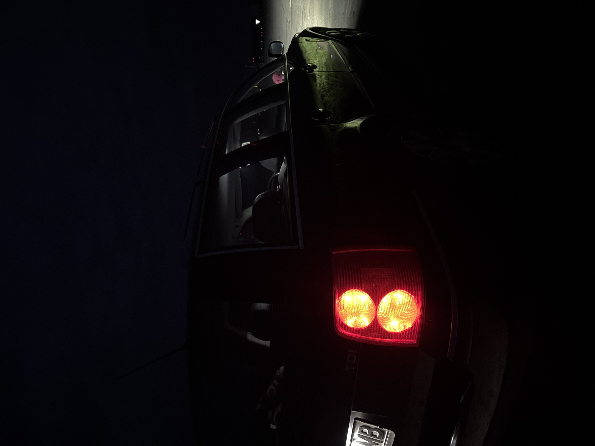 The image depicts a dark-colored car, likely an SUV or hatchback, parked at night in Cherna Gora, Bulgaria. The car is angled diagonally, viewed from a low perspective, showcasing its rear and side. The upper surfaces of the car, including the roof and parts of the side panels, are covered in a textured, greenish-brown material, possibly camouflage, dirt, or moss.
The scene is enveloped in darkness, with the primary light sources being the car's illuminated red taillights, which feature two prominent circular bulbs glowing brightly. A strong, narrow beam of white light shines vertically down onto the upper side of the vehicle, creating a stark contrast and highlighting the car's textured surface against the deep shadows.
Through the rear side window, a glimpse of the car's interior is visible, including seats and a small, pink, possibly plush, object resting on the back seat. Faintly visible on the right taillight is the text "TDI". A portion of the car's license plate is visible at the very bottom, showing the letters "MB". The overall impression is that of a rugged vehicle stopped in a dark, nighttime environment.