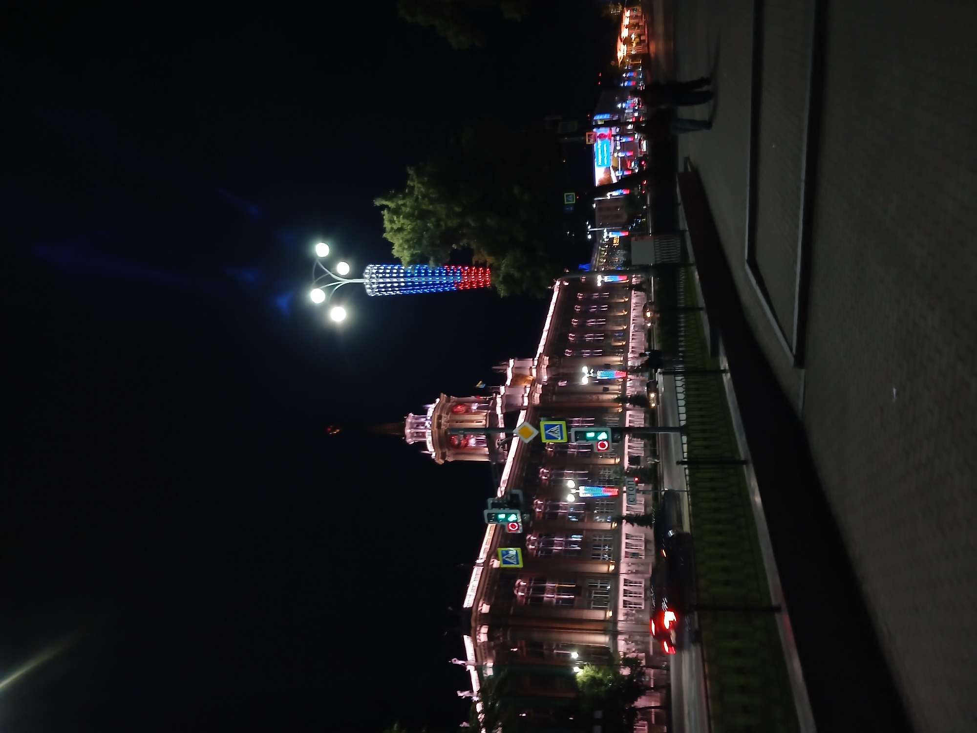 The image depicts a nighttime view of the Yekaterinburg City Hall, also known as the Sevastyanov House, located in Yekaterinburg, Russia. The building is illuminated with warm-toned lights, highlighting its architectural features. In the foreground, a street with passing cars is visible, with traffic lights displaying green signals. A pedestrian walkway lines the street, and two individuals are walking on it in the distance. A tall lamppost decorated with red, white, and blue lights, resembling the Russian flag, stands to the right of the building. The sky is dark, indicating it is night.