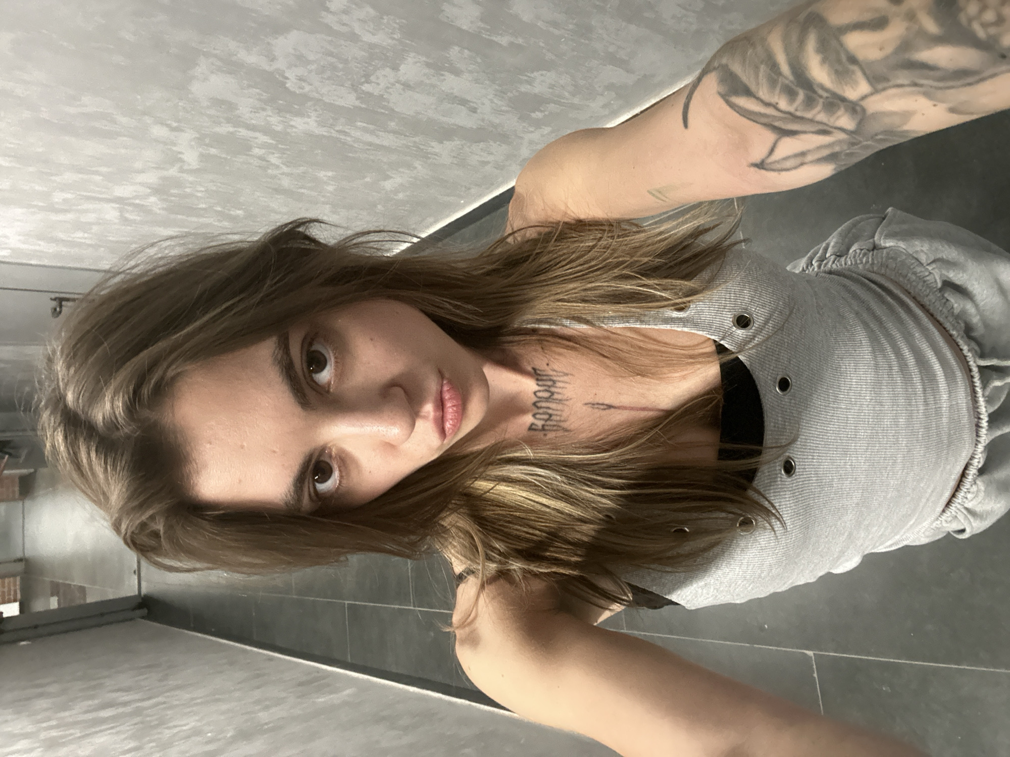A young woman with long, brown, wavy hair, wearing a grey ribbed tank top with grommets and grey sweatpants, stands in a narrow hallway. She has a tattoo on her chest and another on her left arm. The hallway has grey textured walls and a dark tiled floor. The scene is indoors.