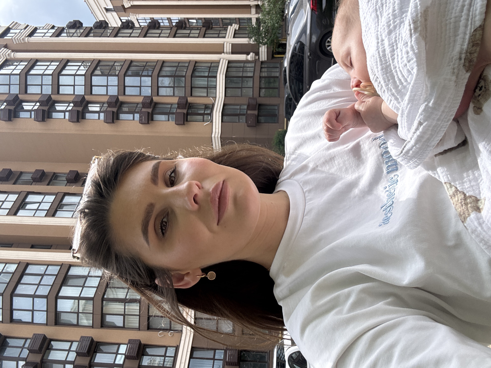 The image depicts a close-up, selfie-style portrait of a woman holding an infant, set in an urban residential area of Sofiivska Borshchahivka, Ukraine. The woman, appearing to be an adult female with brown hair and light skin, is positioned in the foreground, looking directly at the camera. She wears a white t-shirt with the phrase "aesthetic mood" embroidered in light blue script across the chest, and a gold-colored hoop earring with a circular charm is visible on her left ear. Sunglasses are perched on top of her head. In her arms, a swaddled infant, with light skin and closed eyes, has a pacifier in its mouth, with its left hand curled into a fist near its face. The baby is wrapped in a white, textured blanket featuring a subtle pattern, possibly of small bears. The background features several modern, multi-story apartment buildings characterized by their brown and beige facades, numerous large windows (some configured as bay windows with dark frames), and architectural details such as columns and dark-colored planter boxes or small balconies. Multiple dark-colored parked vehicles are discernible in the mid-ground between the subjects and the buildings. The sky is partially cloudy, providing diffused natural lighting for the scene.