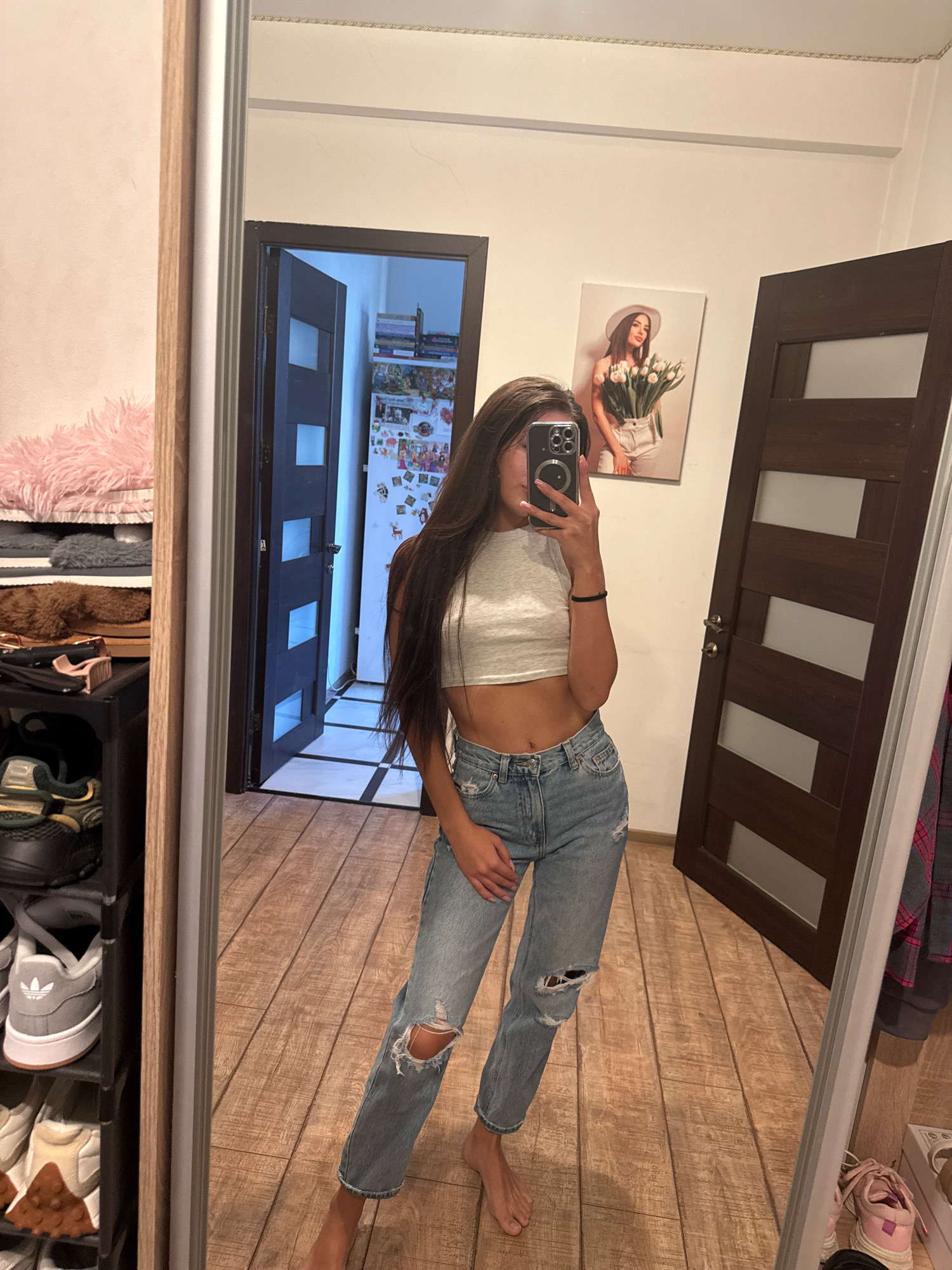 A young woman stands in a room and takes a selfie in a full-length mirror. She has long, dark hair and is wearing a light grey crop top and ripped blue jeans. She is holding a smartphone in her right hand. To her left, there is a tall black shoe rack filled with various pairs of shoes, and a shelf with fluffy pink and grey items. Behind her, a doorway leads into another room which contains a refrigerator covered in magnets and other decorations, and a shelf with books. To her right, a dark wooden door with glass panes stands partially open. On the wall between the doors, there is a framed picture of a woman holding a bouquet of white tulips. The floor is made of wooden planks. The room appears to be a personal living space, possibly an apartment in Chisinau, Moldova.