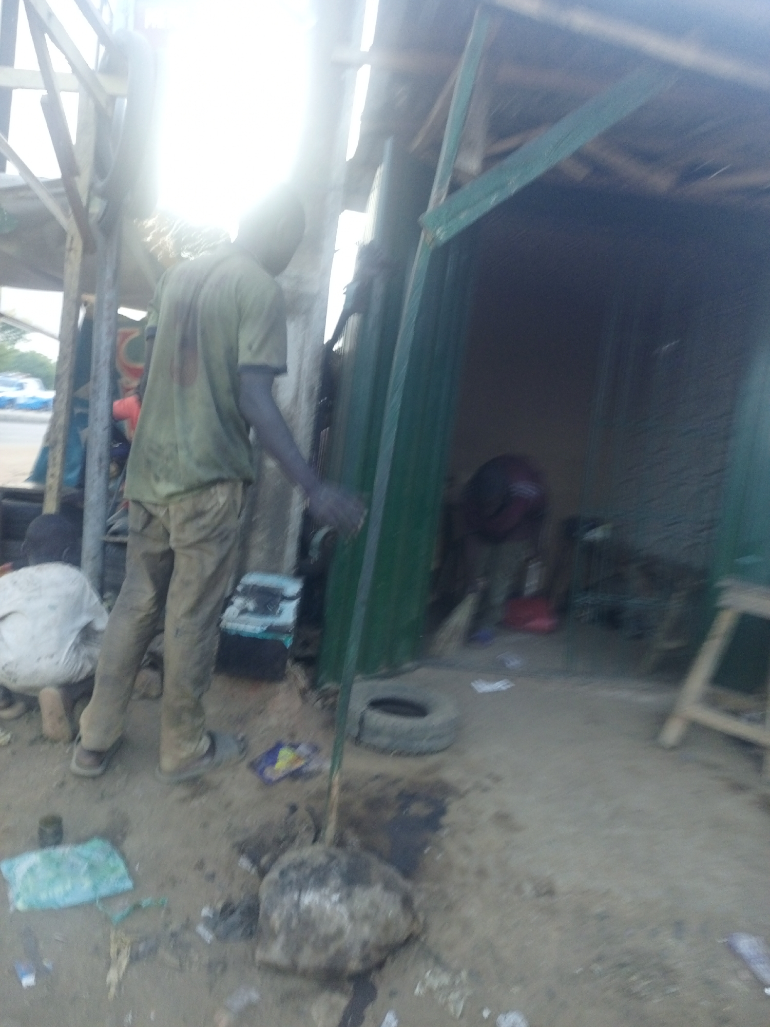 This image captures a vibrant, albeit informal, outdoor workshop scene, likely in Keffi, Nigeria. The setting appears to be a rudimentary structure made of wooden beams and green corrugated metal sheets, serving as a repair shop or stall. The ground is unpaved, covered in dirt and scattered debris, including plastic bags, a rock, and what appears to be a small tire.
In the foreground, a man is visible from behind, wearing a dirty light green short-sleeved t-shirt and light brown trousers. His arms appear to be covered in a dark, greasy substance, suggesting he is engaged in manual labor, possibly mechanical work. His stance indicates he is either actively working or momentarily paused, facing into the shaded area of the structure.
To the lower left, another individual is partially visible, crouched down and wearing a white or light-colored garment, also appearing to be involved in an activity on the ground. Inside the shaded area of the workshop, various tools and indistinct objects are present, including what might be a fan or piece of machinery, further supporting the idea of a repair or craft space.
The time of day is bright, with strong sunlight streaming from the upper left, causing significant glare and overexposure in that section of the image. The sun casts highlights and shadows, but the overall brightness suggests a daytime setting, possibly late morning or early afternoon. The outdoor background is blurry but shows hints of vehicles or other structures in the distance. No specific text is clearly visible in the image, and emotions are not discernible given the subjects' postures and distance. The scene conveys a sense of a bustling, hands-on work environment.