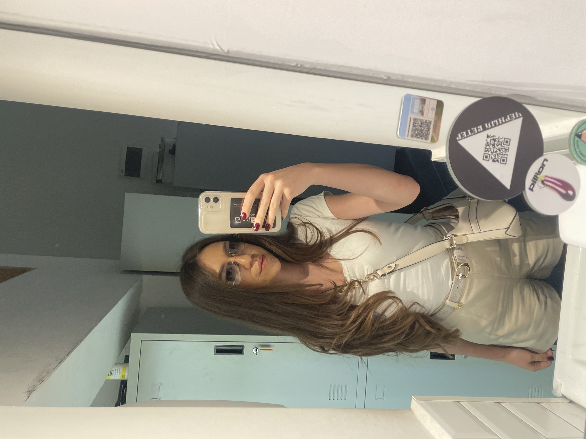 The image shows a woman taking a selfie in a mirror. She is wearing sunglasses, a white top, and light-colored pants. She has long brown hair. The background reveals an interior setting, with a metallic storage cabinet and white walls. The presence of stickers on the mirror featuring Cyrillic script ("Черный Ветер") suggests the location may be in a country using the Cyrillic alphabet. Given the "pillion" sticker, the location is likely Sofia, Bulgaria, as "pillion" is a popular brand in Sofia.