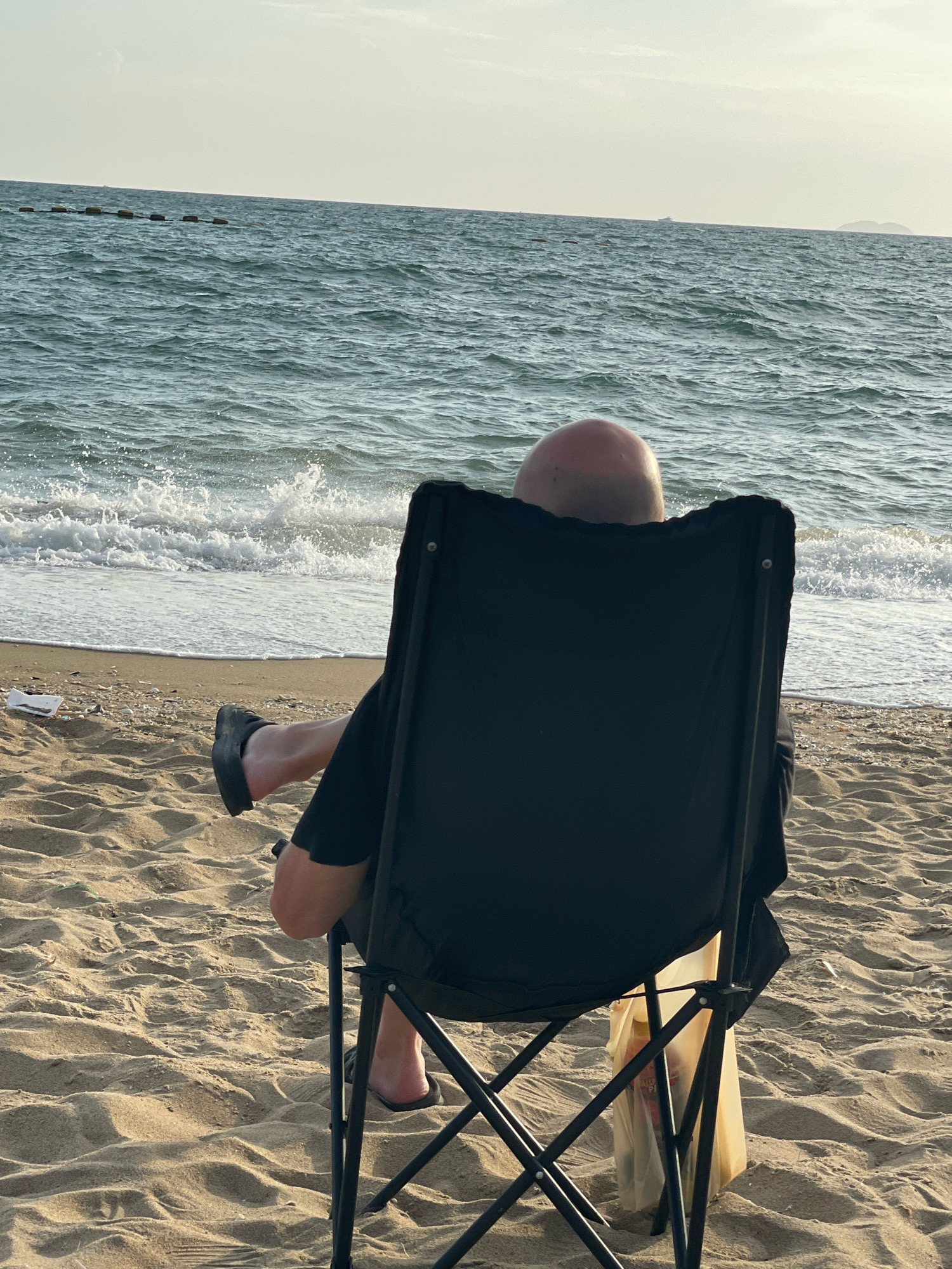 A lone, bald individual is seated in a black folding chair facing the ocean on a sandy beach in Pattaya City, Thailand. The person is wearing dark clothing and flip-flops. Gentle waves are breaking on the shore. The sky is overcast with a pale yellow hue, suggesting either early morning or late afternoon. The background shows a line of buoys in the water and a distant island or landmass. The beach is sparsely populated, with a plastic bag hanging from the chair.