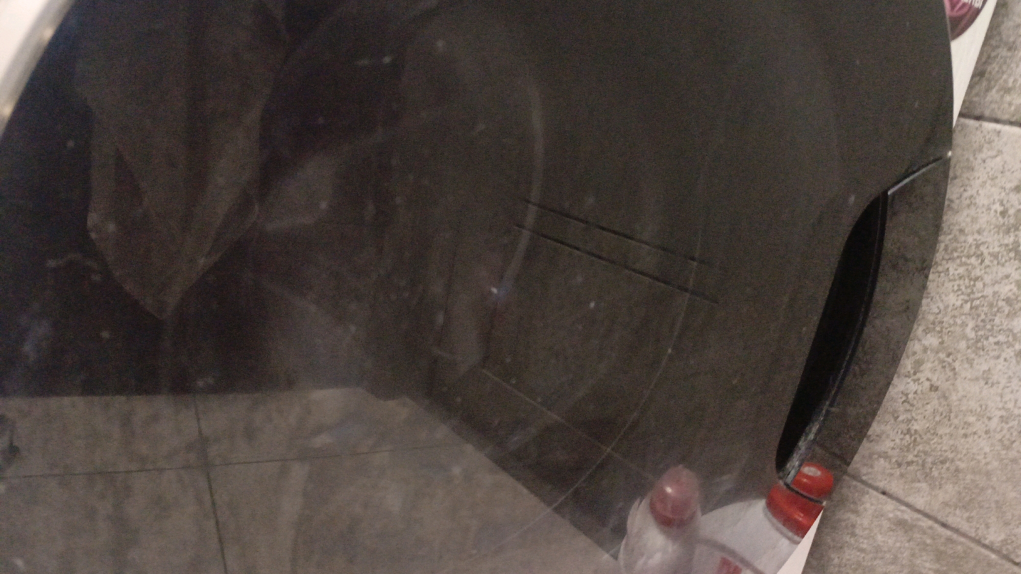 The image shows a close-up view of a dark, reflective surface, possibly a countertop or appliance. There are two faint horizontal lines etched into the surface. In the bottom right corner, a white plastic bottle with a red cap is partially visible, and to its left, another bottle with a pinkish cap is also visible. The background appears to be tiled flooring. A reflection on the dark surface suggests the presence of objects and perhaps a person or persons, but these are indistinct. The scene is likely indoors. Without further information or context, it is not possible to determine the specific location within Homyel, Belarus.