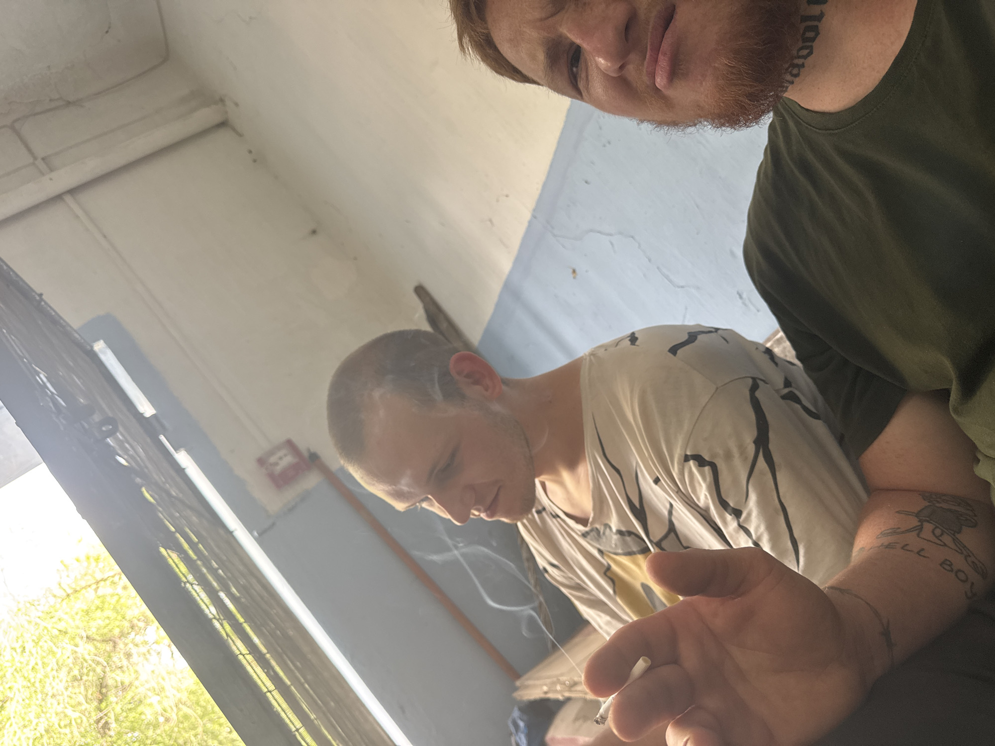 Two men are outdoors, possibly in an urban setting, given the concrete and metal structures visible. The man on the left, with a shaved head and light stubble, is holding a cigarette and exhaling smoke, his gaze directed to the left. He is wearing a white t-shirt with black lines. The man on the right, with red hair and beard, is facing the camera with a slightly grimacing expression and is wearing a dark green t-shirt. He has a tattoo on his neck that appears to say "maboll". Both men have tattoos on their arms. The background shows a blue wall and overhead structure with what appears to be sunlight filtering through, suggesting it might be daytime. There's a fire alarm on the wall. The overall impression is a candid, informal moment.