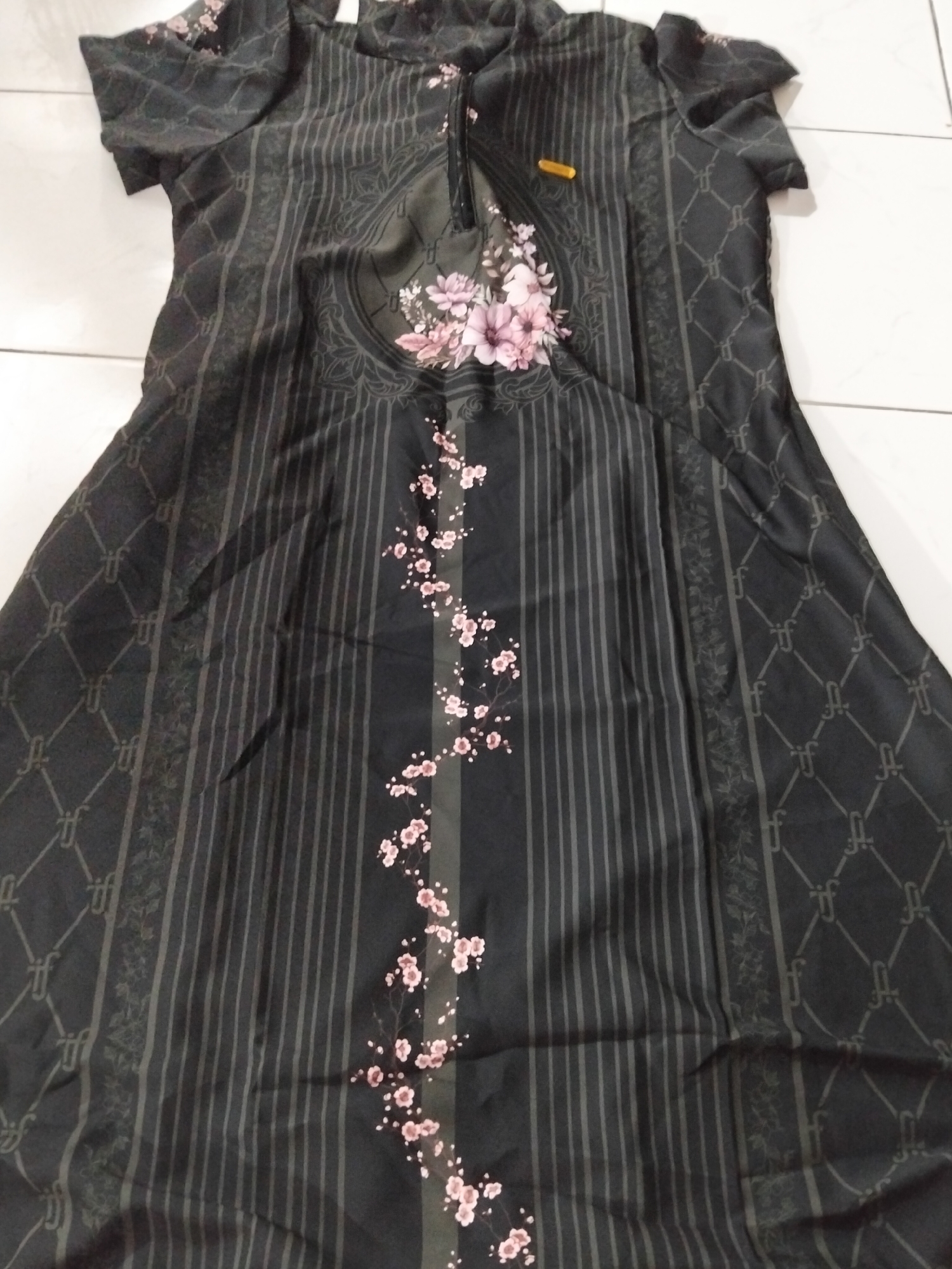 A black dress with a floral pattern is laid out on a white tiled floor. The dress has short sleeves and a high neckline with a zipper. The pattern features vertical stripes, a diamond-shaped lattice, and scattered pink flowers. The dress is oriented vertically, with the top part towards the top of the image. The lighting is somewhat dim, creating shadows on the fabric. The scene appears to be indoors, possibly in a bedroom or a display area, given the tiled floor. There is no indication of the dress being in Payagaboh, Indonesia, as this information is not present in the image.