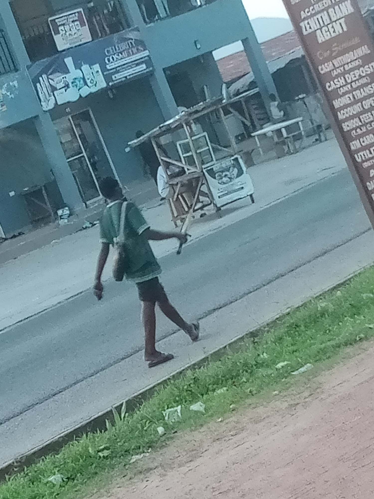 The image shows a street scene in Jalingo, Nigeria. In the foreground, a young male wearing a green t-shirt and dark shorts is walking on the right side of a paved road. He has a bag slung over his right shoulder and is holding a stick or similar object in his right hand. To the right of the road, there is a strip of green grass with some litter. In the background, there is a building with a sign that reads "CELEBRITY COSMETICS" and another sign for "ZENITH BANK AGENT". There are also some makeshift stalls and a vehicle visible in the background. The overall environment appears to be an urban or semi-urban setting with moderate activity.