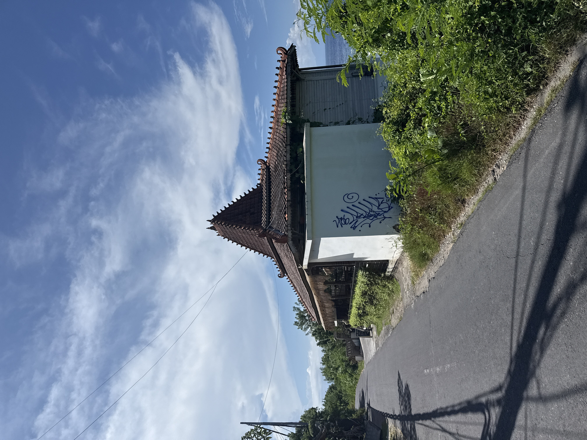 The image shows a street scene in Batununggal, Indonesia. A single-lane asphalt road runs along the right edge of the frame, with tree shadow projected across it. A building with a tiered, orange-tiled roof is located on the left side of the road. The building has a white exterior wall with blue graffiti. Lush green vegetation borders the road and building. The sky is visible in the upper left portion of the frame, featuring scattered clouds.