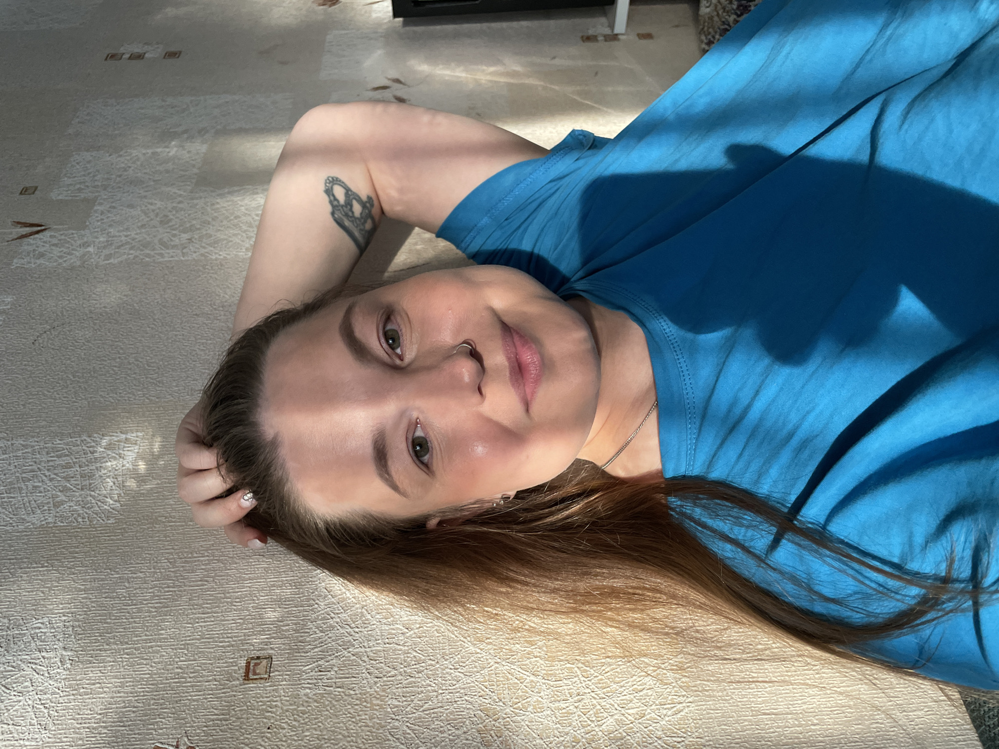 A woman is depicted lying on her back on a light-colored, textured floor or carpet, looking directly at the camera with a relaxed, slightly smiling expression. Her head is positioned towards the upper right of the frame, and her body extends diagonally towards the lower left.
The woman has long, light brown hair spread around her head and flowing down her back. She has green-brown eyes and a small silver hoop nose ring in her right nostril. She is wearing a vibrant blue short-sleeved t-shirt and a delicate silver necklace. A black and white tattoo, resembling a crown or an ornate butterfly, is visible on her left upper arm. Her left hand is gently placed near her head, with light-colored polish and a small design on one fingernail.
The scene is indoors, likely a domestic setting, with strong natural sunlight casting distinct patterns of light and shadow across the floor and her t-shirt, indicating a sunny daytime. No specific location cues are visible within the image itself beyond the provided context of Izhevsk, Russia. There is no visible text.