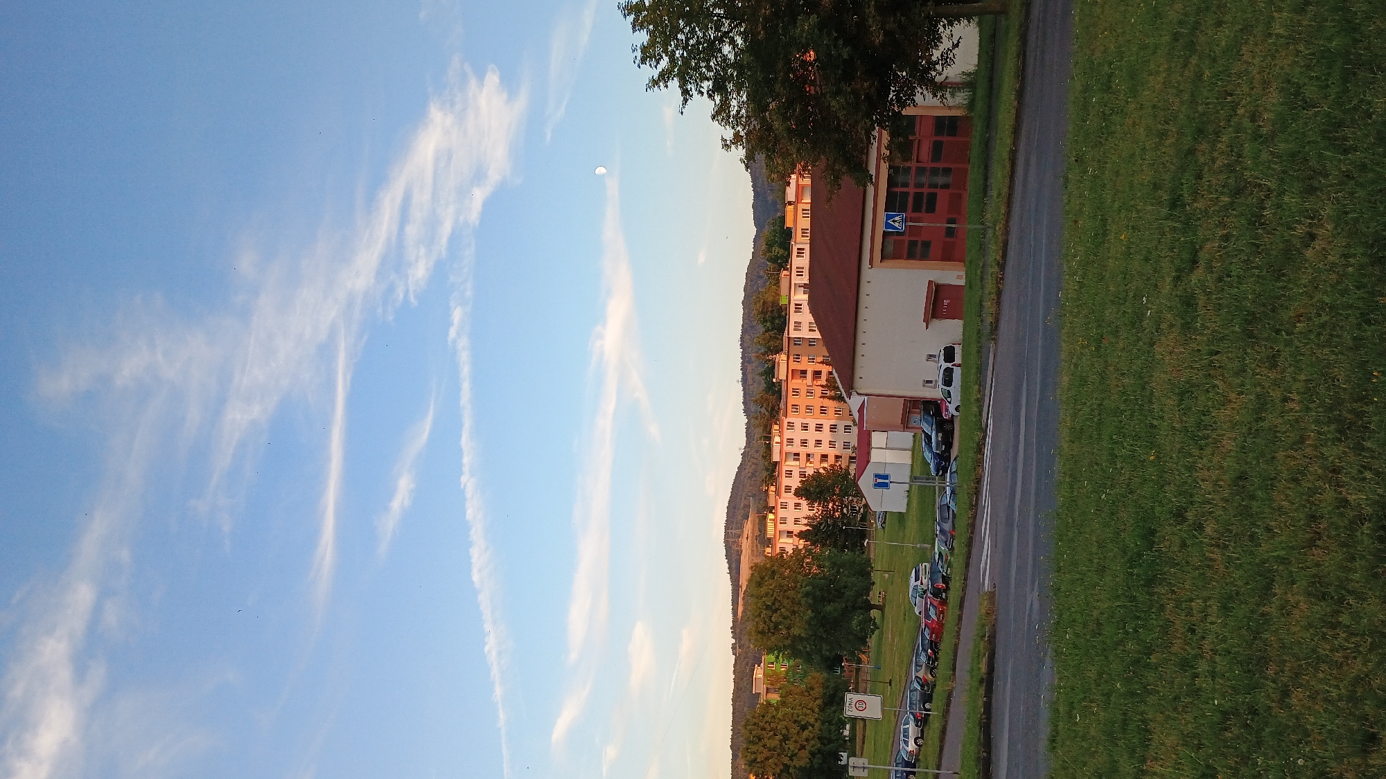 The image captures an outdoor scene in Sokolov, Czechia, likely during the late afternoon or early evening, given the warm lighting and the visible moon in the sky. The foreground features a grassy embankment sloping down towards a road. Beyond the road, a parking lot is filled with numerous cars of various colors, including silver, black, red, and blue. A speed limit sign with "30" is visible on the left side of the parking lot.
Further back, several buildings are present. The most prominent is a long, multi-story residential building with a light-colored facade and numerous windows, its balconies adorned with plants. To its right, a smaller, single-story building with a red roof and garage-style doors is situated, possibly a fire station or similar facility, indicated by a blue and white pedestrian crossing sign in front of it. A larger, reddish-brown building with a red roof is also visible. Behind these structures, a densely wooded hill forms the backdrop, its slopes covered in dark green trees. The sky is a clear blue, interspersed with wispy white clouds, and the moon is a pale crescent shape. The overall impression is of a suburban or residential area within a town.