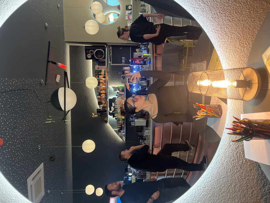 This image captures a mirror selfie taken inside a bar or cafe in Rafailovići, Montenegro. The primary subject, a female with dark hair, eyeglasses, and a dark long-sleeved top, is reflected centrally, holding a smartphone with a visible multi-lens camera array to capture the image. Her head is turned slightly to her left.
The circular mirror has a textured, light-colored frame. In the immediate foreground, on a dark table, sit a black mesh lamp with a visible warm-glowing filament bulb, a black mesh container filled with colored pencils, and a white piece of paper.
The reflected bar interior features a dark, speckled ceiling with multiple spherical pendant lights and a prominent Saturn-like pendant light with a black ring. An air conditioning vent and a red wall-mounted object are also visible on the ceiling. The walls are predominantly dark grey/black, textured, with a section of lighter, possibly beige, wall on the right featuring circular decorative elements.
Behind the bar counter, a male with a beard and dark clothing stands on the left, hands in his pockets. Another male in dark clothing leans on the bar in the center-left, with one foot on a rail. On the right, a female in dark clothing stands behind the bar, facing away from the mirror, with her hands on her lower back.
The bar itself has an illuminated, reddish-pink front panel. Behind it, multiple shelves are stocked with various bottles of alcohol, glassware, a coffee machine, and other bar equipment. A golden statue or trophy is visible on the top shelf. A black speaker cabinet is also positioned on a shelf. A yellow chair or stool is partially visible behind the bar.