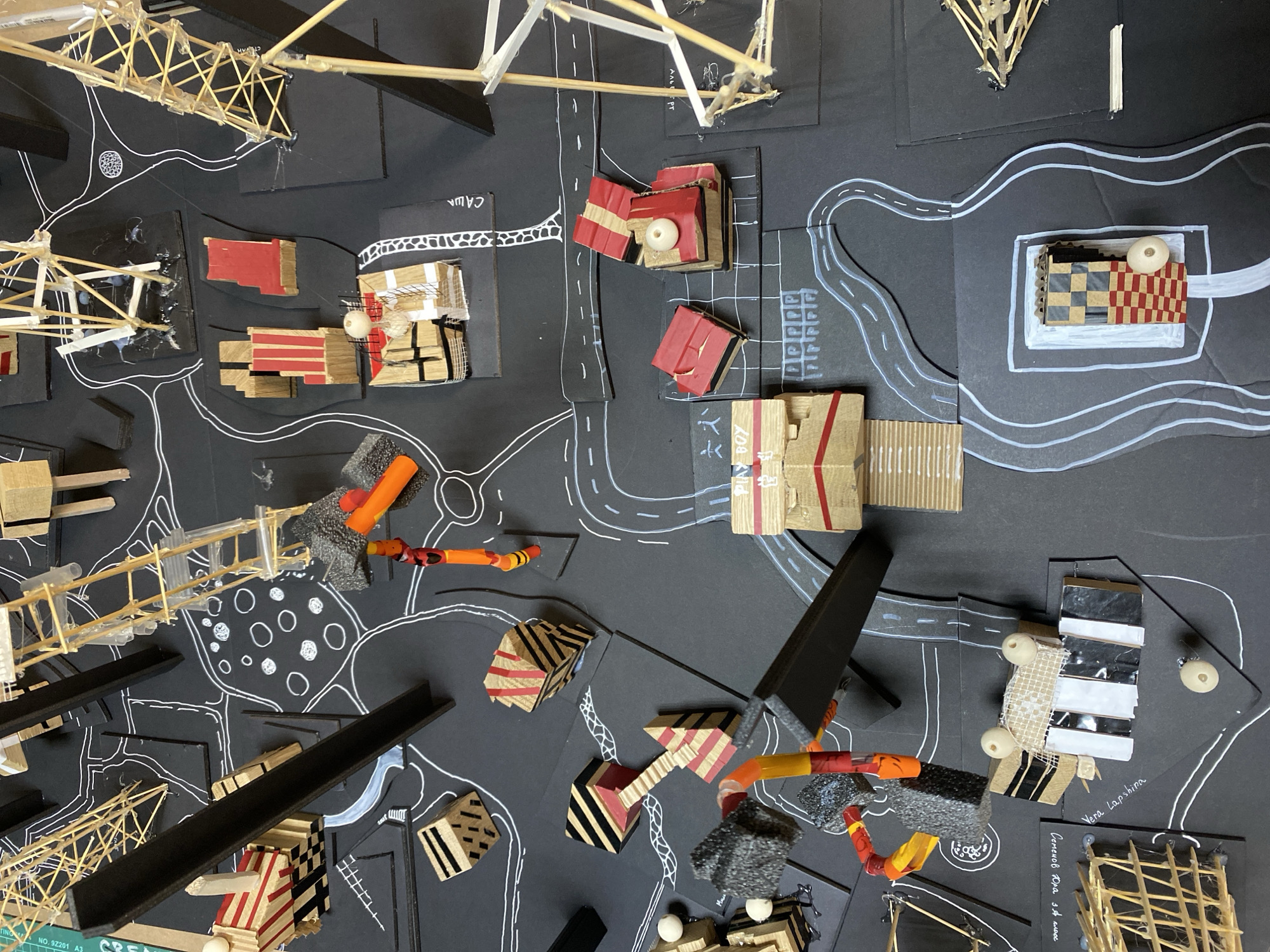 This is a high-angle view of a model city layout on a black background. The model includes roads drawn with white lines, various building structures made of cardboard, wood, and other materials. Some structures are painted or decorated with red stripes and other patterns. There are also abstract representations of trees or greenery. Certain areas are labeled with handwritten text in what appears to be Russian. Specifically, I can see the name "Vera Lapshina," and "Семенов Юра за класс." I also see "PLAY BOY," and "CAWA." It is difficult to ascertain if this is a specific depiction of Moscow.