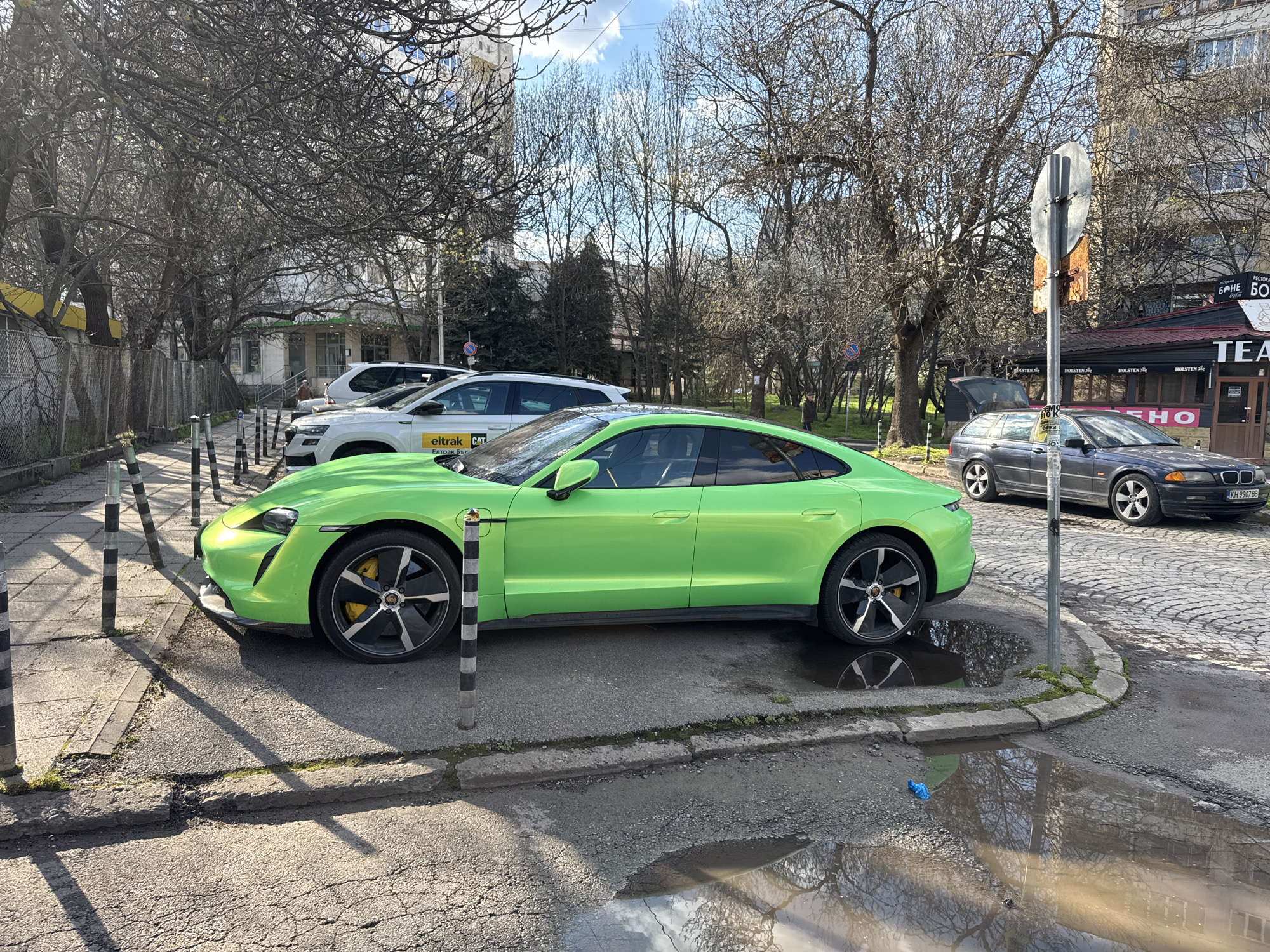 The image shows a green Porsche Taycan parked on a street corner in Sofia, Bulgaria. The car is positioned between black and white striped bollards and a puddle reflecting the bare branches of nearby trees and the sky. Behind the Taycan, there are other parked vehicles, including a white SUV with the "eltrak CAT" logo and a dark gray BMW station wagon with license plate "KH 9907 BB". A street sign and a building with a sign reading "TEA" are also visible in the background. The sidewalk is cracked and the street appears to be cobblestone or paved with stone blocks. The sky is partly cloudy, suggesting it is either late fall, winter or early spring.