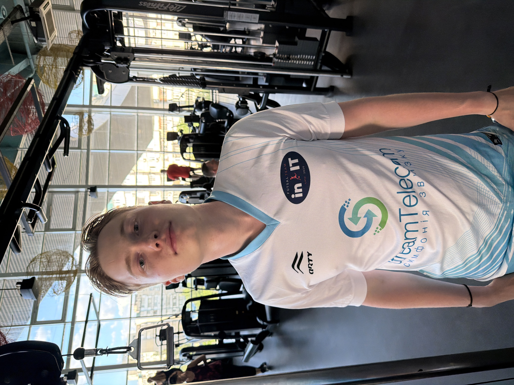A young, fair-skinned man with light brown hair stands in a gym, looking directly at the camera. He is wearing a white t-shirt with blue accents, featuring logos and text. The t-shirt has a V-neck collar and short sleeves. On the left chest area of the t-shirt, there is a black logo with wavy lines and the word "ARTY" below it. The right side of the chest has an oval logo with text that reads "FUTSAL TEAM in IT LVIV 2020". Below that, there is a large graphic with a stylized "S" made of green and blue arrows. The lower portion of the t-shirt has a pattern of light blue stripes and text in Ukrainian. He has a black bracelet on his left wrist and an orange string bracelet on his right wrist. The man's expression is neutral.
The background of the image reveals a gym environment. Various weight training machines are visible, including cable machines and multi-gyms. In the distance, other gym-goers can be seen, some using equipment. The gym has large windows that let in natural light, and decorative elements, possibly made of straw or wicker, hang from the ceiling. The floor is dark-colored, likely rubber or similar gym flooring. The overall atmosphere suggests an indoor fitness facility.