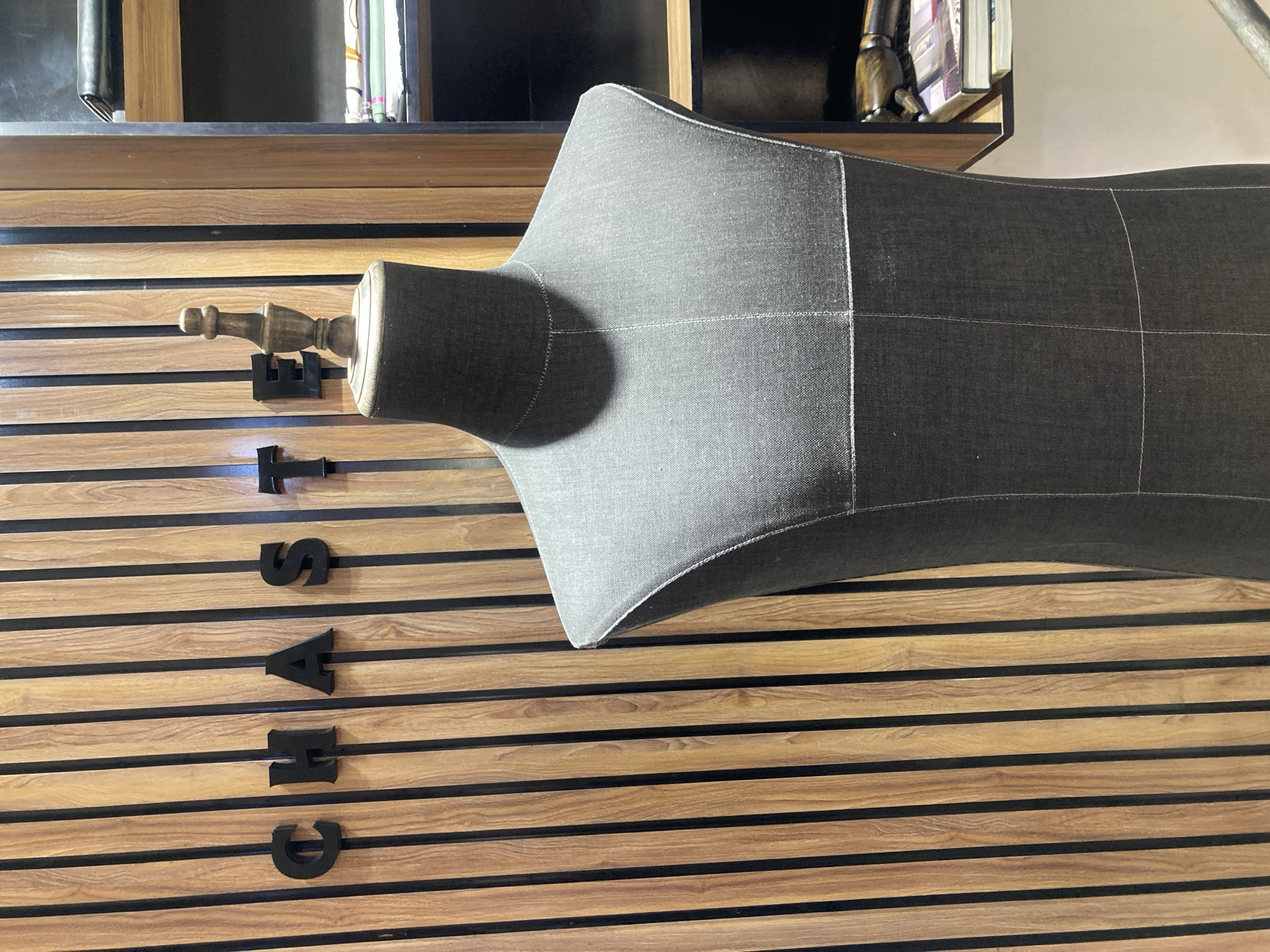 A sideways, eye-level shot captures a dress form mannequin positioned on a wooden wall with horizontal brown planks separated by black lines. The mannequin is gray and sectioned off with white seams. Attached to the neck is a wooden knob-like piece. Vertically spelled out to the left of the mannequin are the black capital letters "CHARTSTAND". The wooden wall is below a shelf with books and other objects on it. The setting is likely a shop or studio in Jos, Nigeria. There are no people visible.