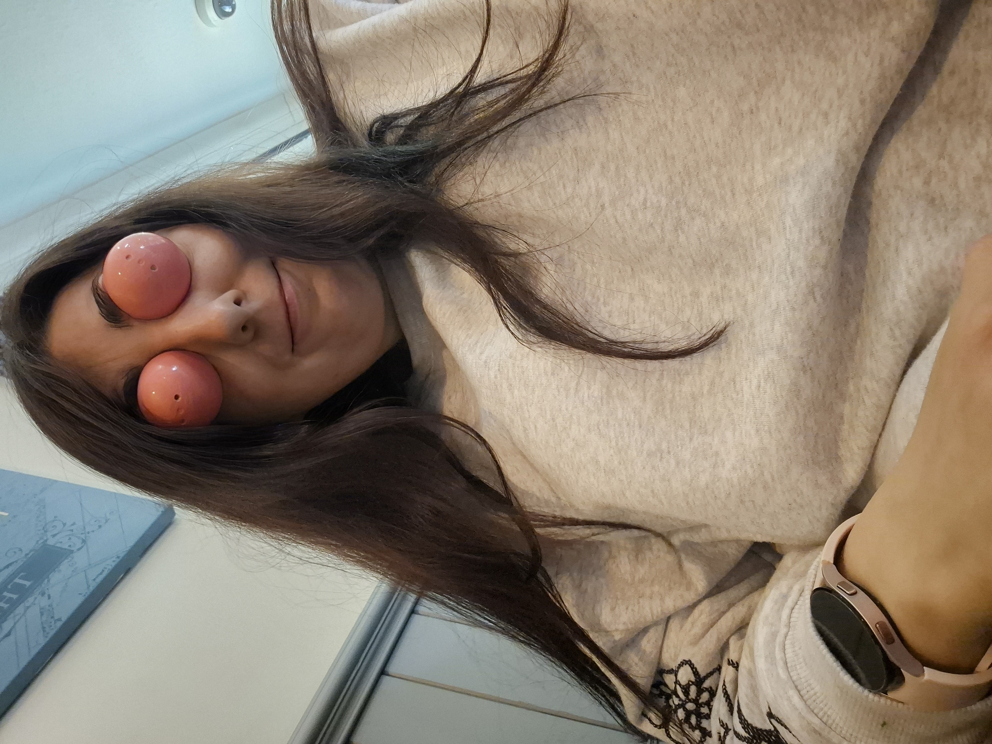 Here is a description of the image:
A woman with long, dark hair is wearing a light-colored hooded sweatshirt. Her eyes are covered with pink spherical objects with small holes. She is smiling slightly. She is wearing a smartwatch with a pink band on her wrist. The background includes a blue object with white lettering, a white wall, and a ceiling with a light fixture. The location cannot be determined as Breckland District, United Kingdom from visual information.